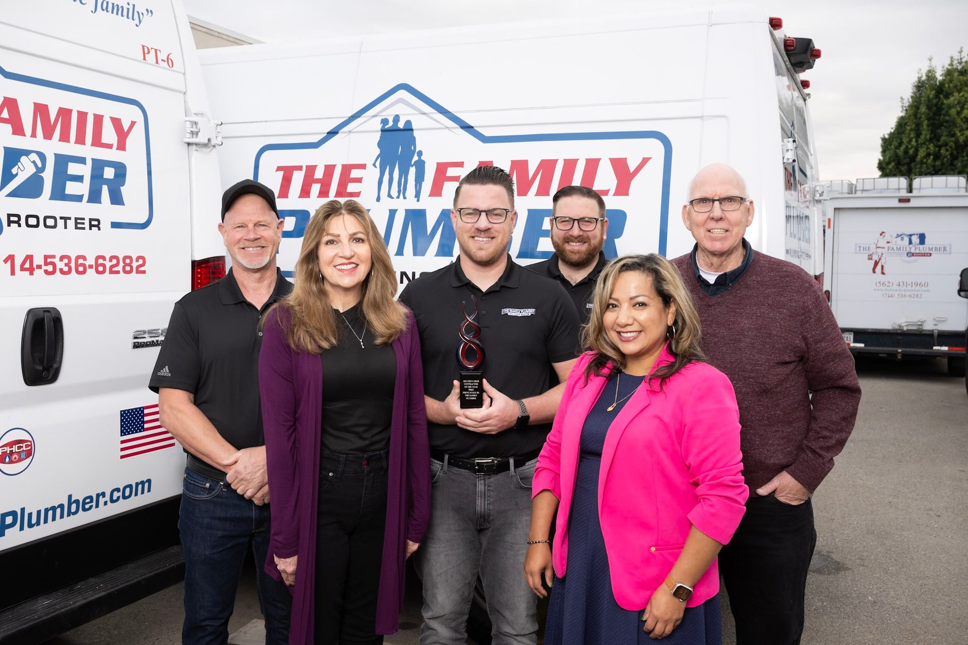 Six people, including one holding an award, stand in front of "The Family Plumber" vans.