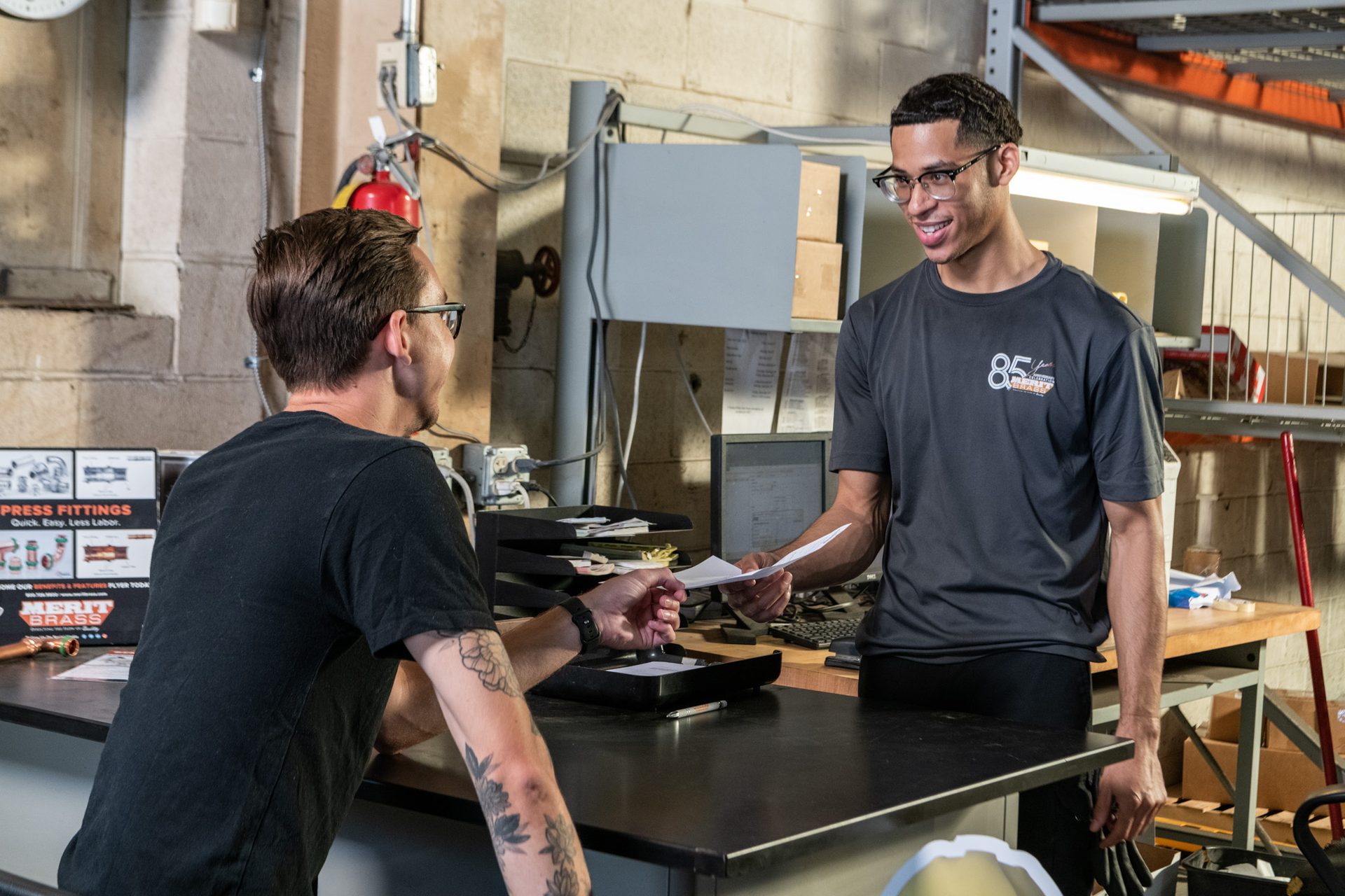 Two smiling men exchanging papers at a counter in a workshop or warehouse.