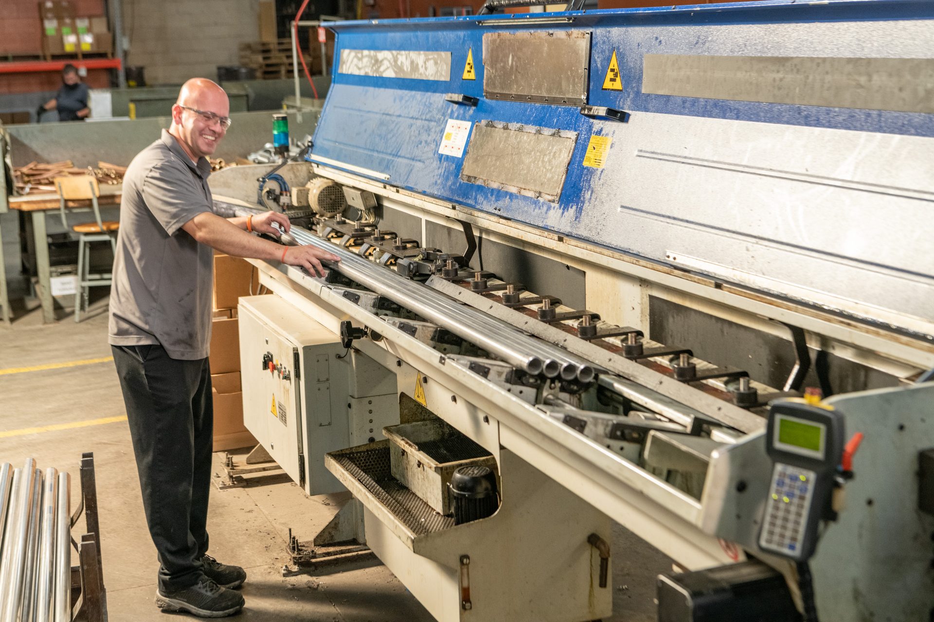 Man smiling, operating a blue industrial machine with metal tubes in a factory.