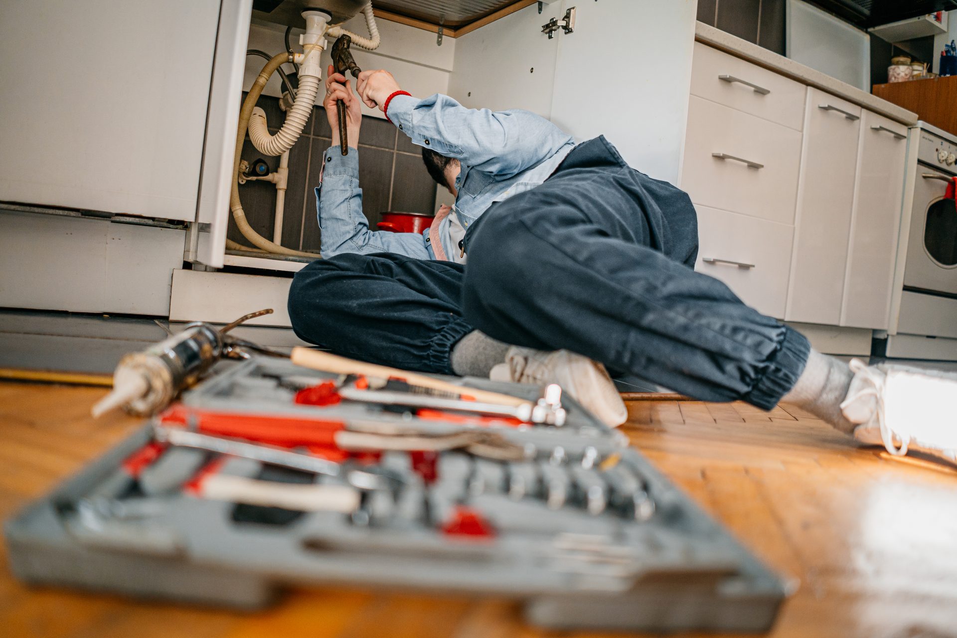 A person lies on the floor, repairing pipes under a kitchen sink with a toolbox and tools nearby.