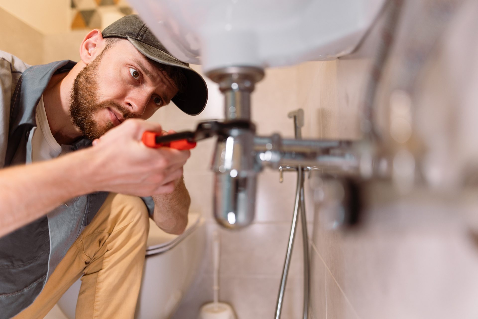 A plumber wearing a cap and beard is fixing a sink drain with a wrench in a bathroom.