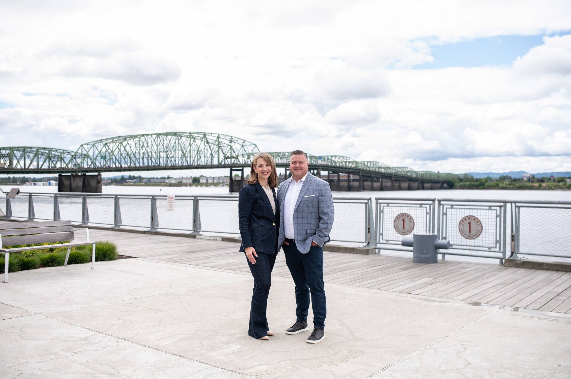 A man and a woman smiling, standing outdoors by a river with a green truss bridge in the background.