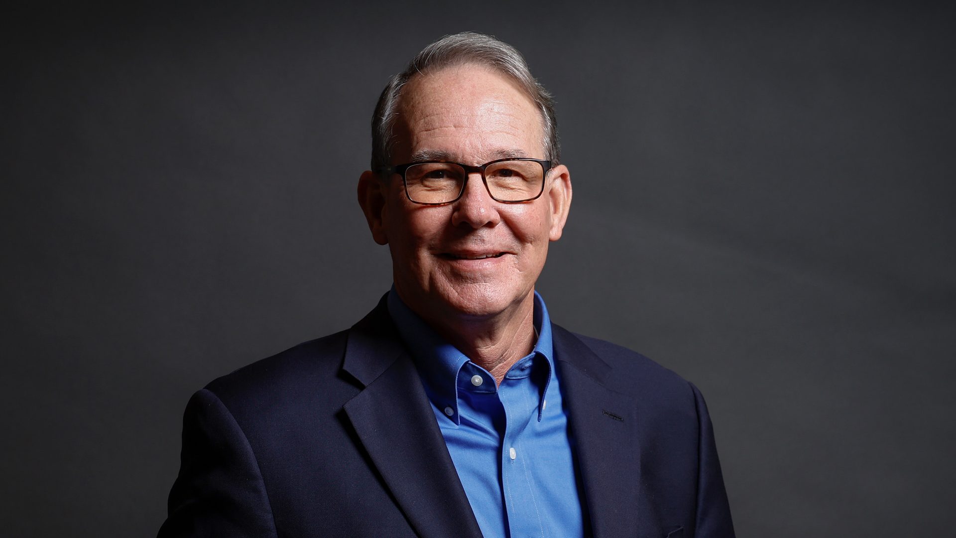 Smiling man with glasses, blue shirt, dark blazer.