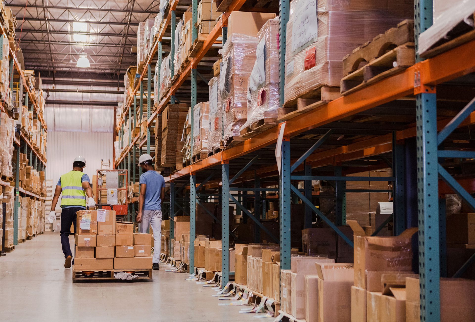 Two workers move boxes on a pallet jack in a busy warehouse.