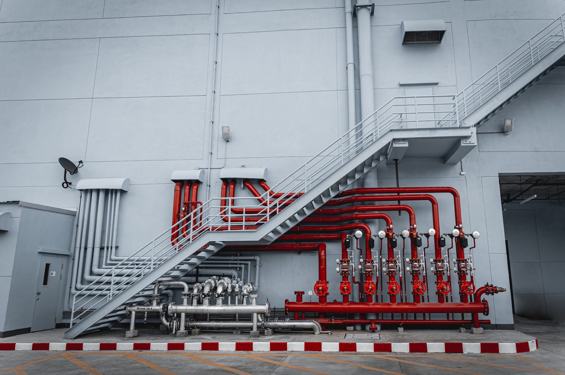 Gray industrial building exterior with a metal staircase and a complex system of red and gray pipes.