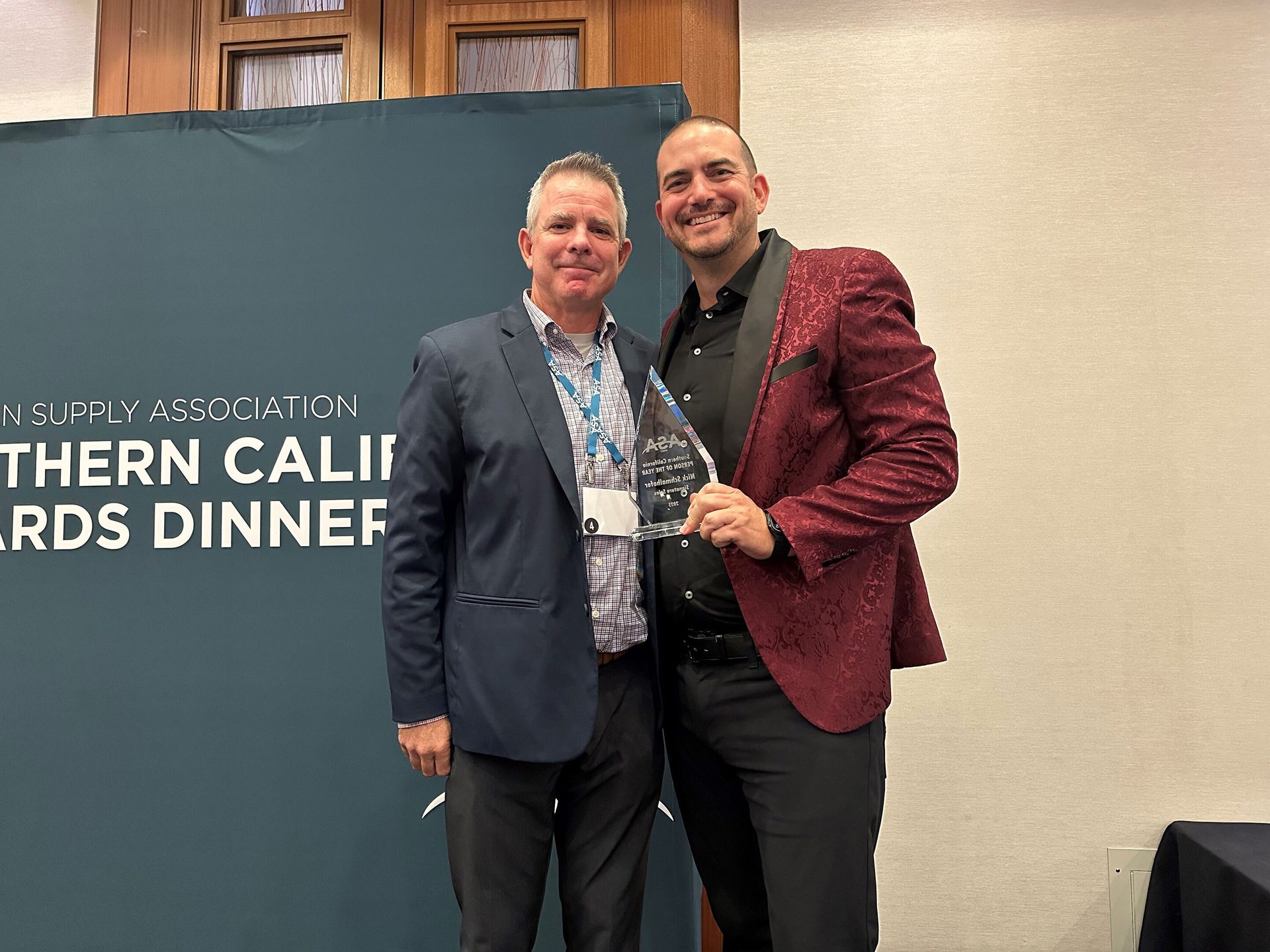 Two men, one in a red patterned blazer, holding a 'Person of the Year' award at a dinner event.