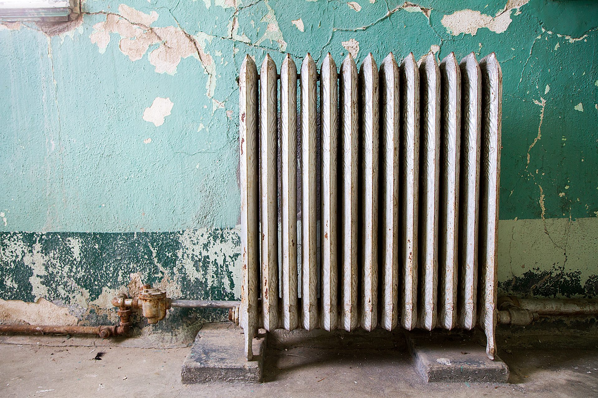 An old white radiator stands against a peeling turquoise and green wall with rusted pipes.
