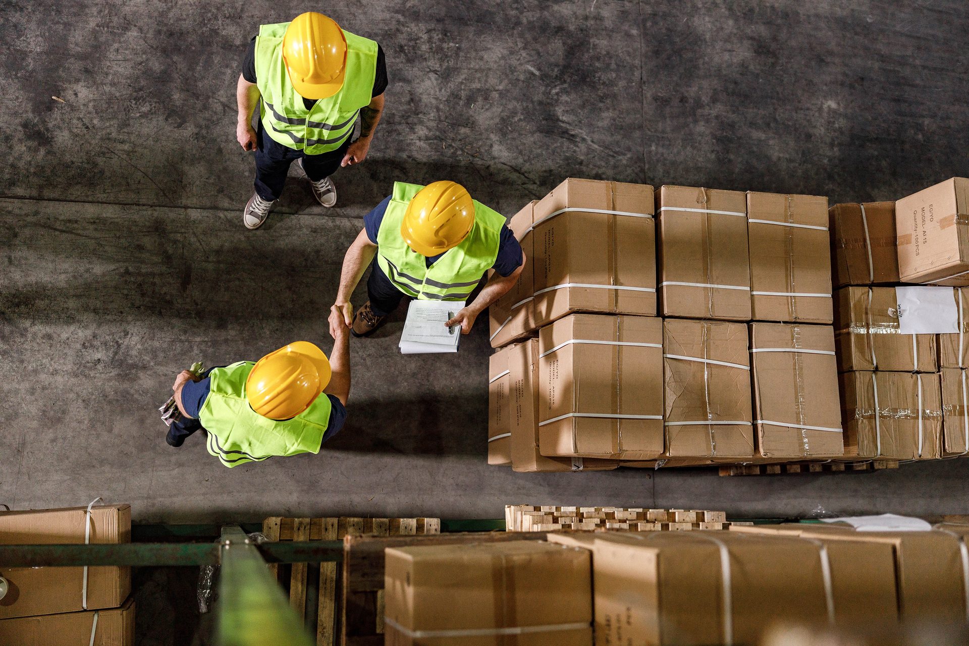 Overhead view: Three workers in hard hats and vests, two shaking hands near stacked boxes in a warehouse.