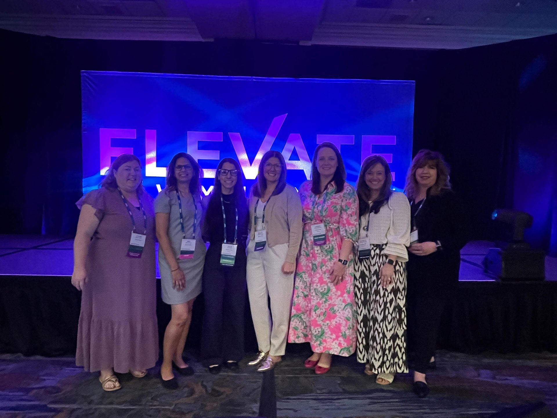 Seven women standing on stage in front of a blue "ELEVATE" banner.