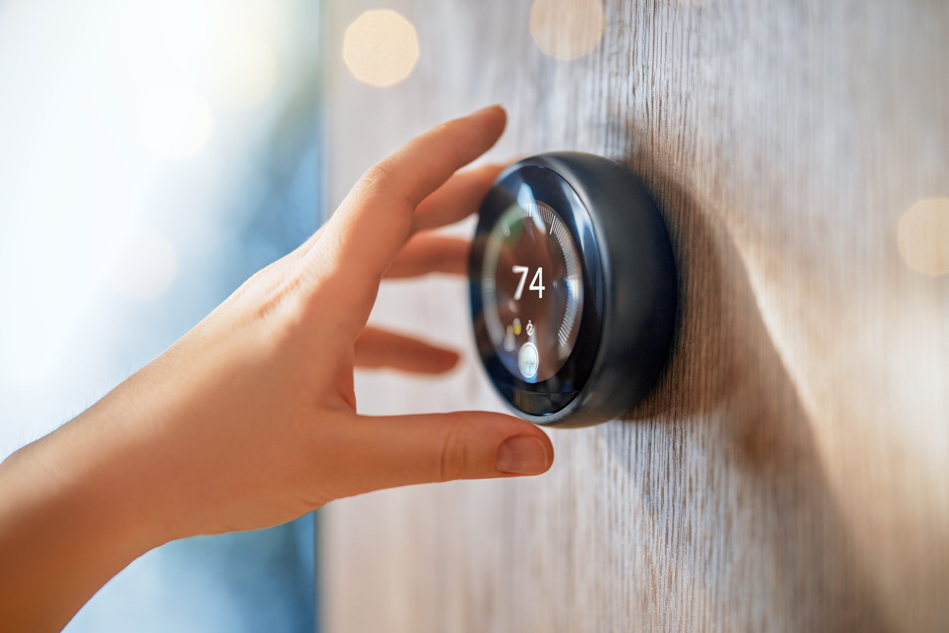 Hand adjusting a smart thermostat displaying 74 degrees on a wooden wall, indicating home climate control.
