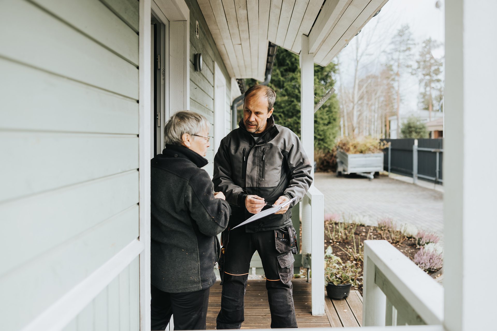 A man in work clothes and an older woman discuss documents on a house porch, likely a contractor meeting a client.