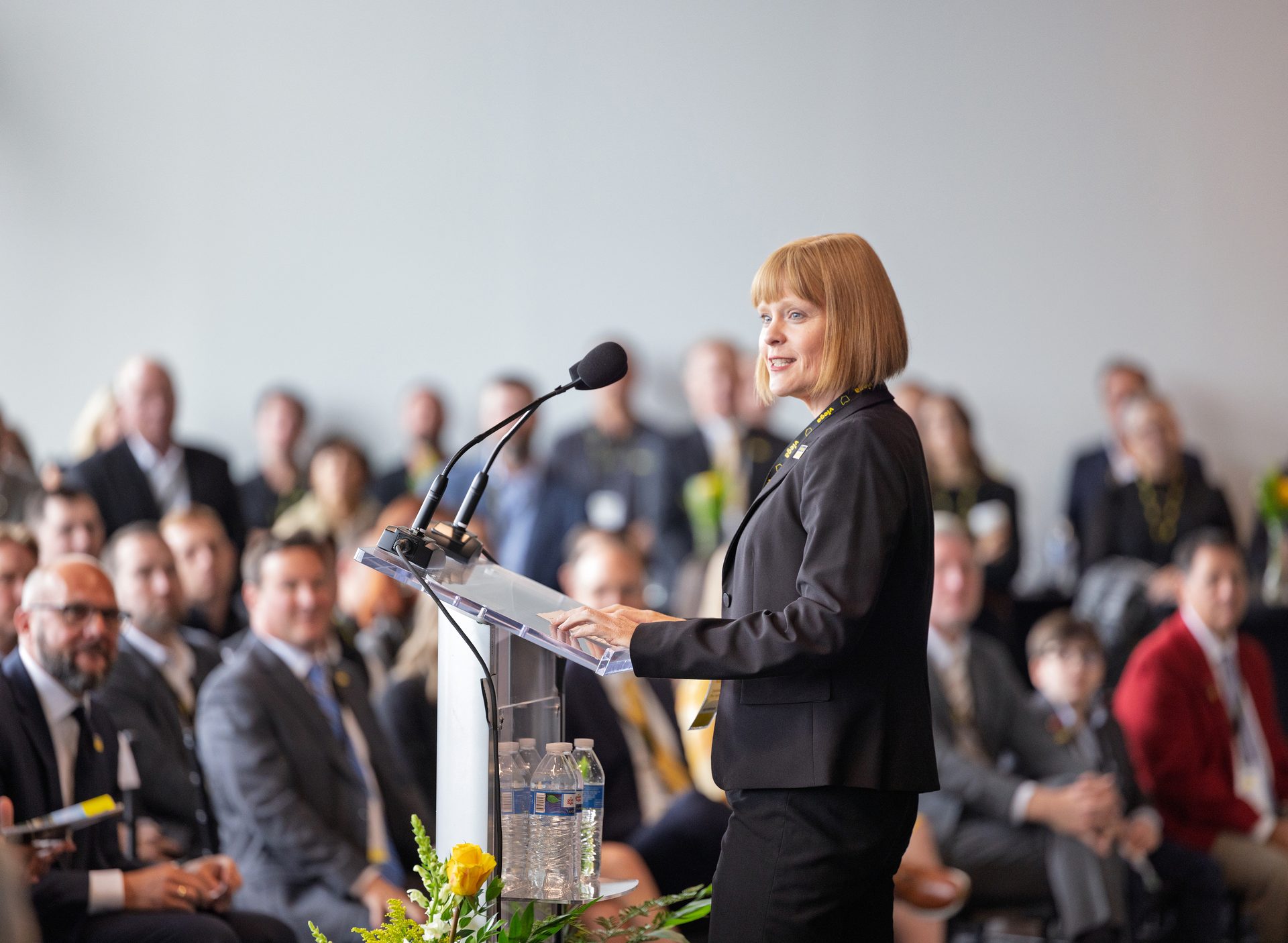 Woman with short blonde hair speaking at a clear podium with microphones to an audience in a bright room.