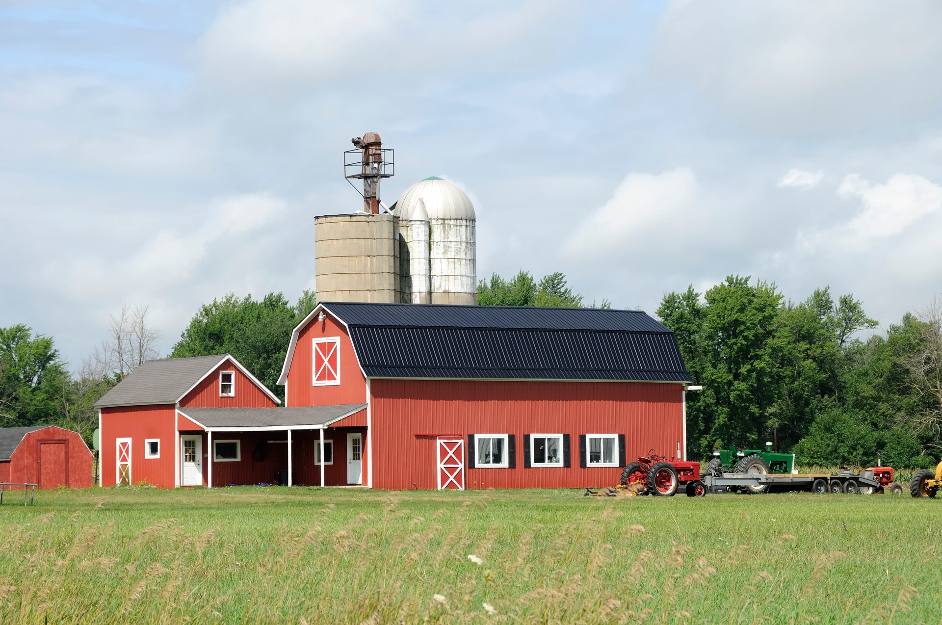 Farm with red barn, silos, and tractors in a green field under a cloudy sky.
