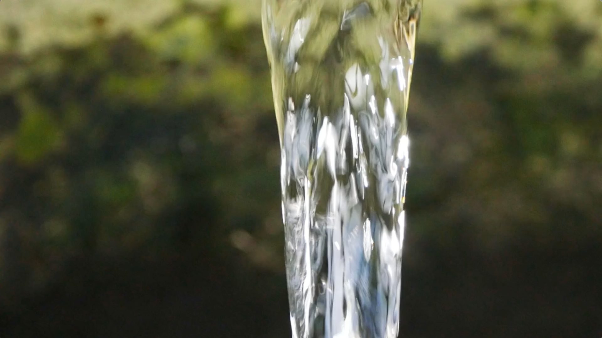 A clear stream of water flows vertically against a blurred green and dark background.