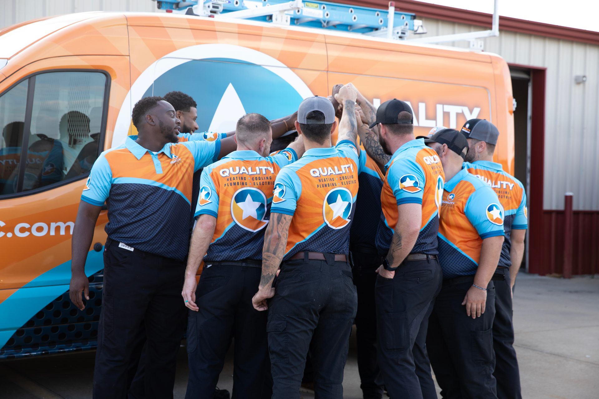 A team of service technicians in orange and blue uniforms huddle together in front of their company van.