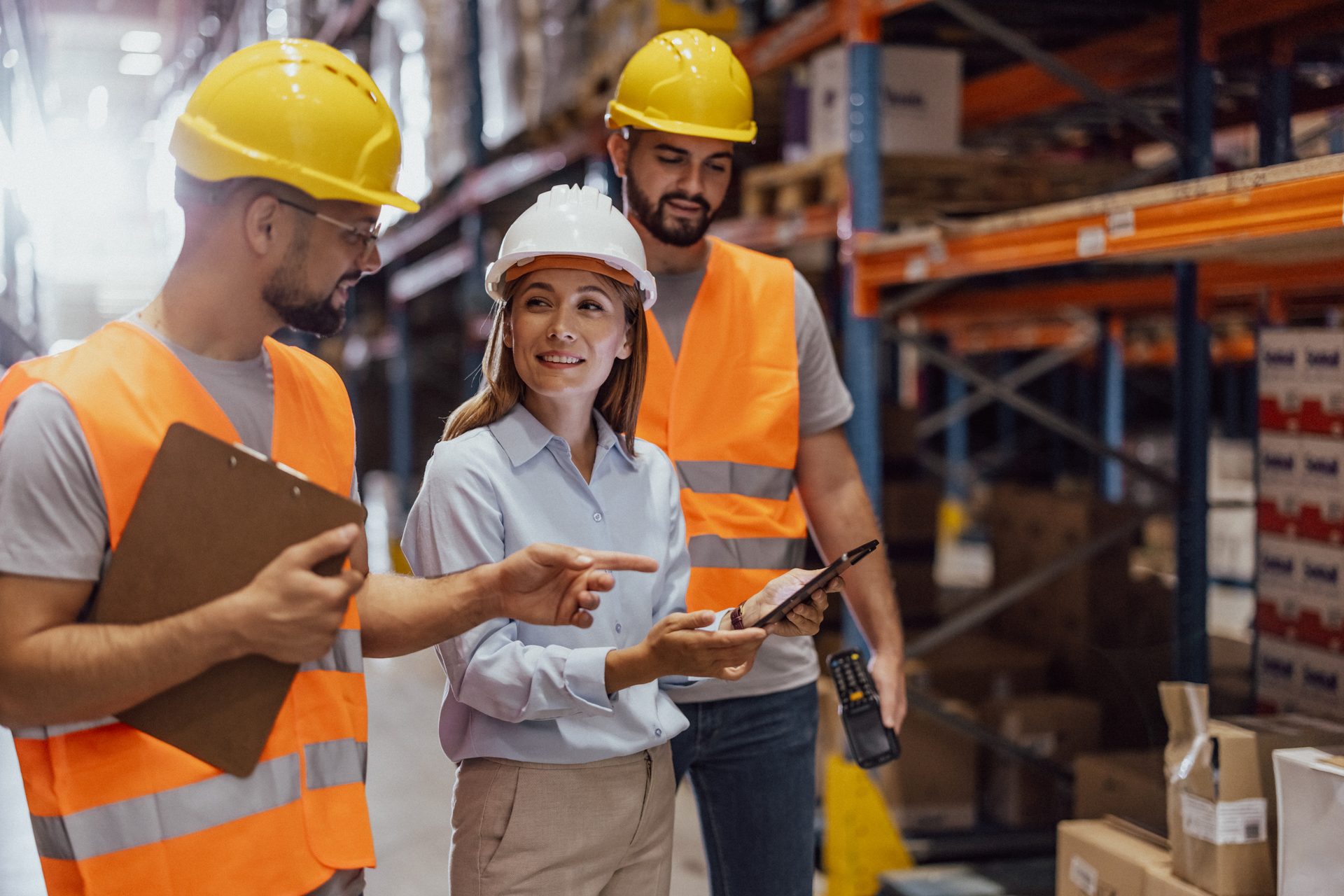 Three warehouse workers in safety gear with a tablet and clipboard.