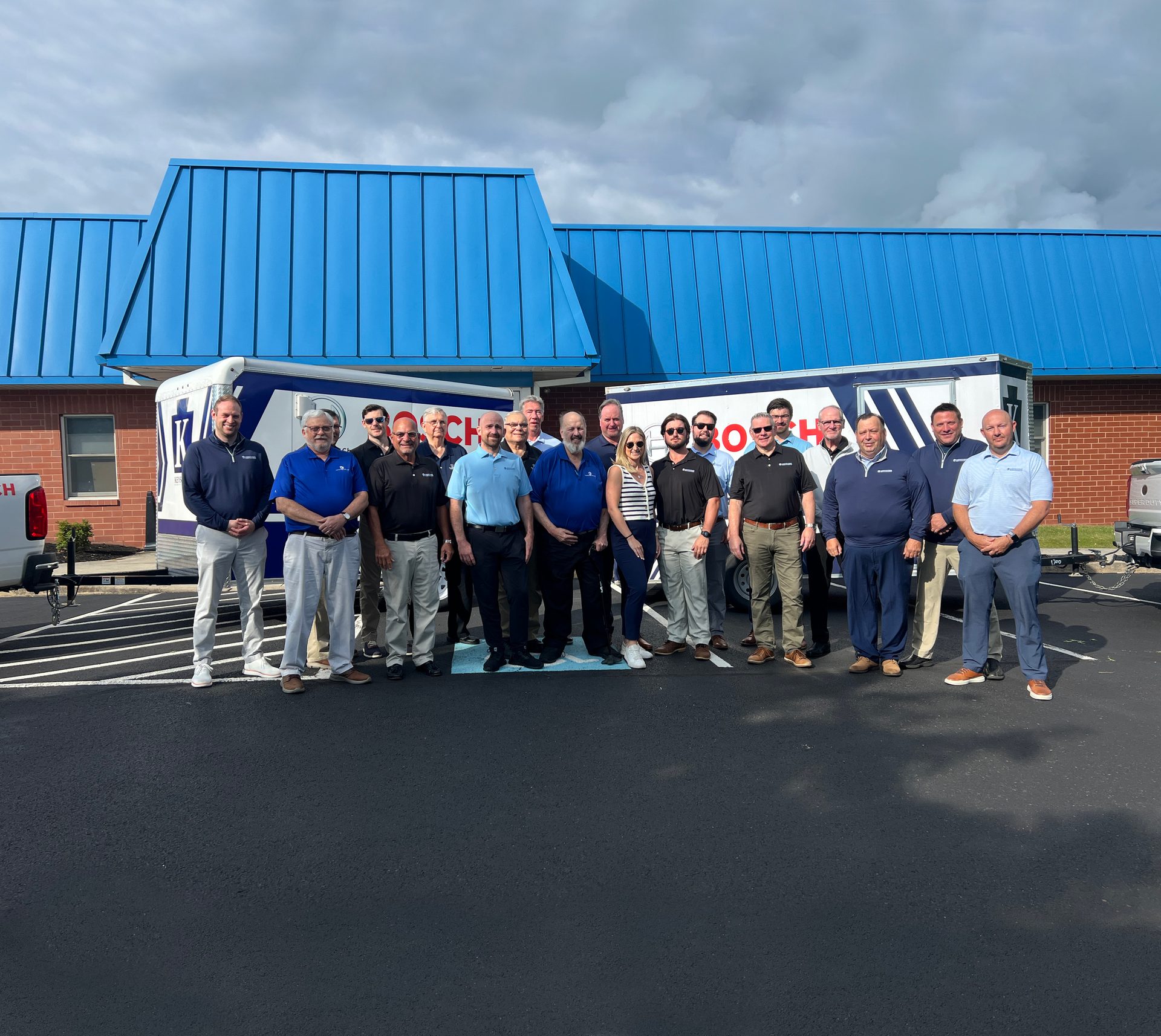 A group of professionals, mostly men in blue shirts, standing in front of two trailers and a building.