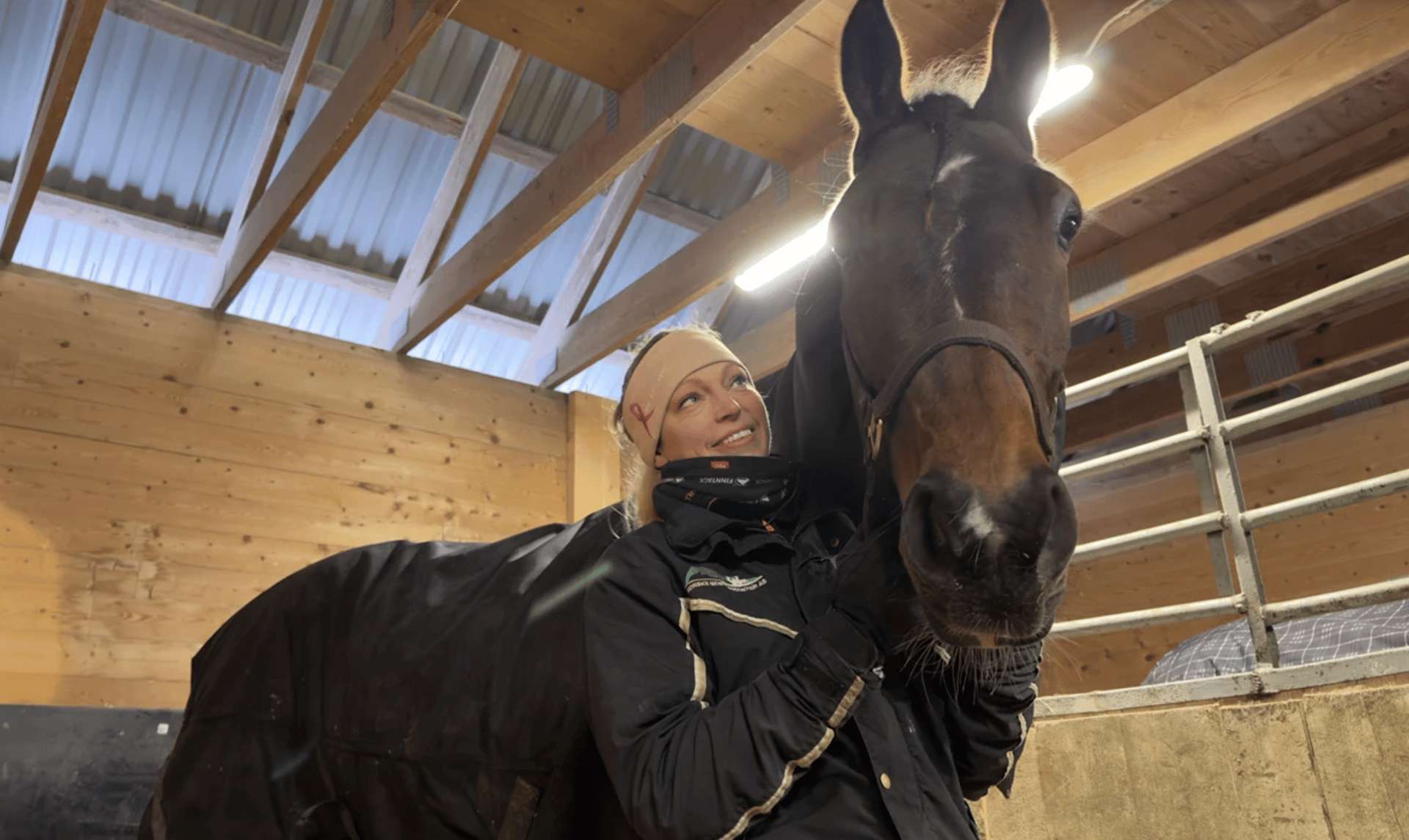 A woman in a stable smiles up at a horse with a blanket on, both in a warm, rustic setting.