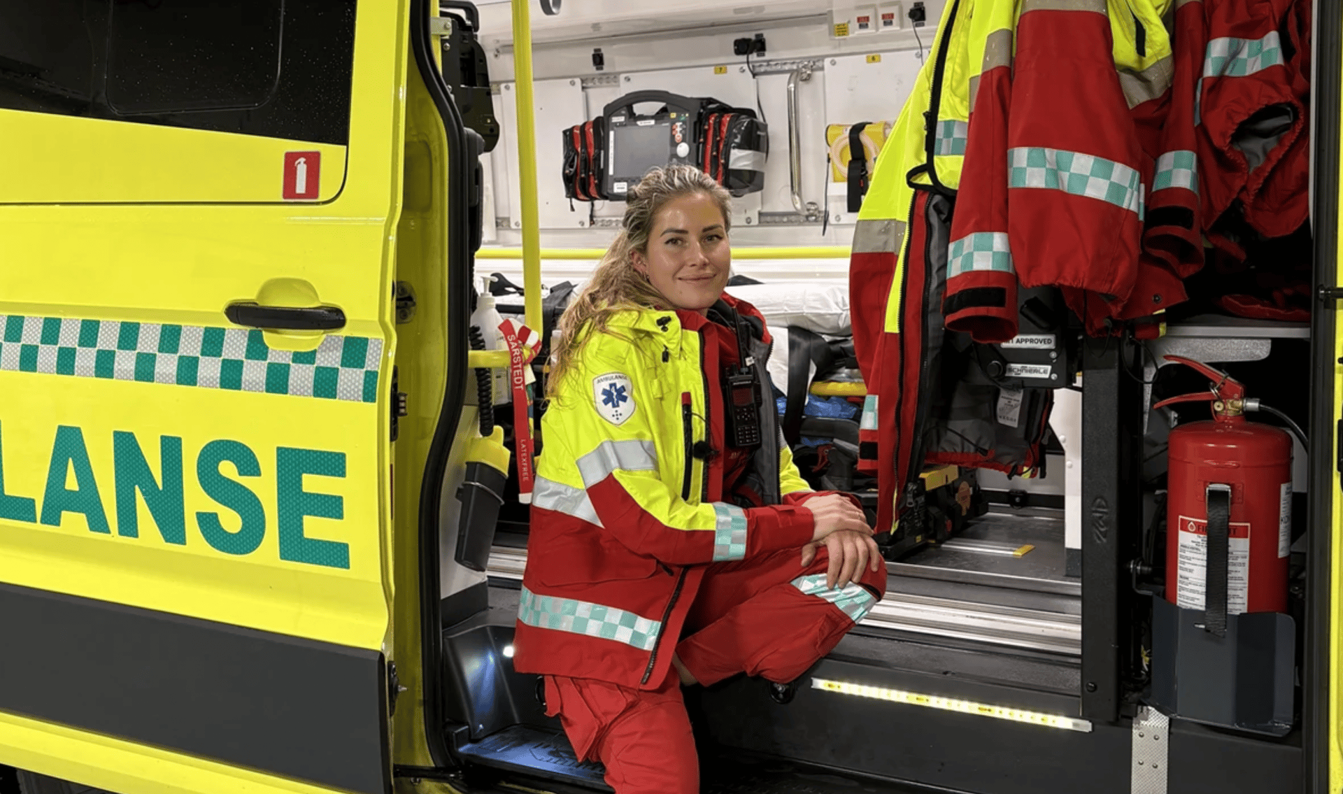 A female paramedic in a red and yellow uniform crouches inside a yellow ambulance, smiling.