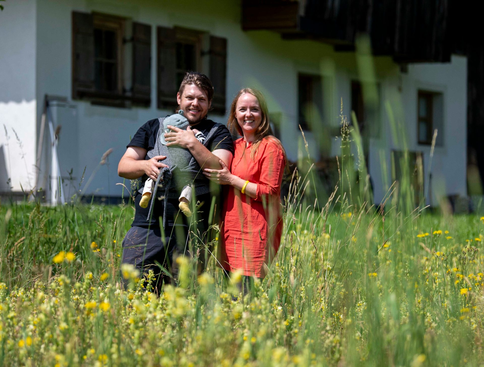Smiling couple holding a baby in a carrier, standing in a flowery field with a house.