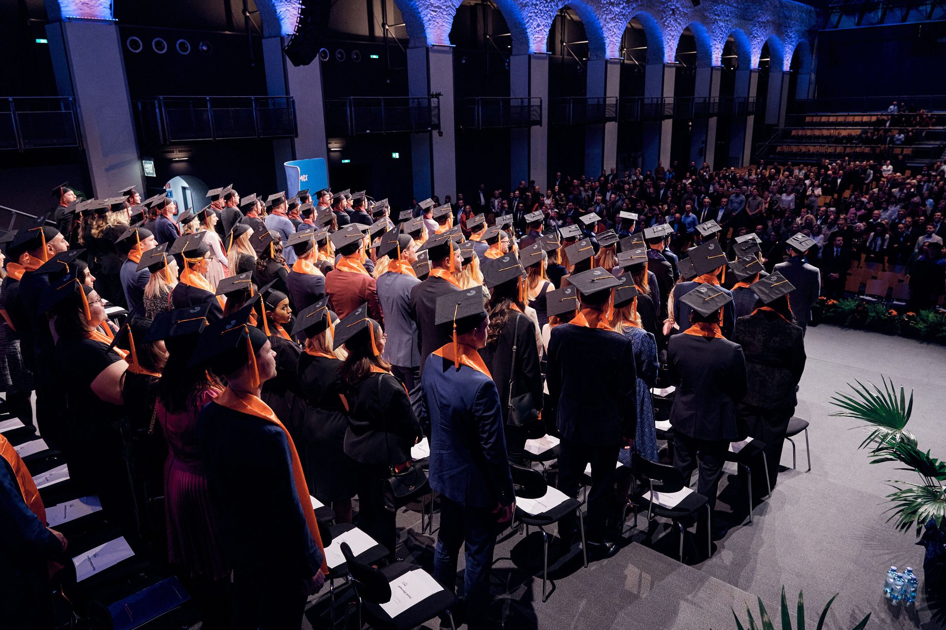 View from behind of numerous graduates in black caps and orange stoles at a large indoor ceremony.