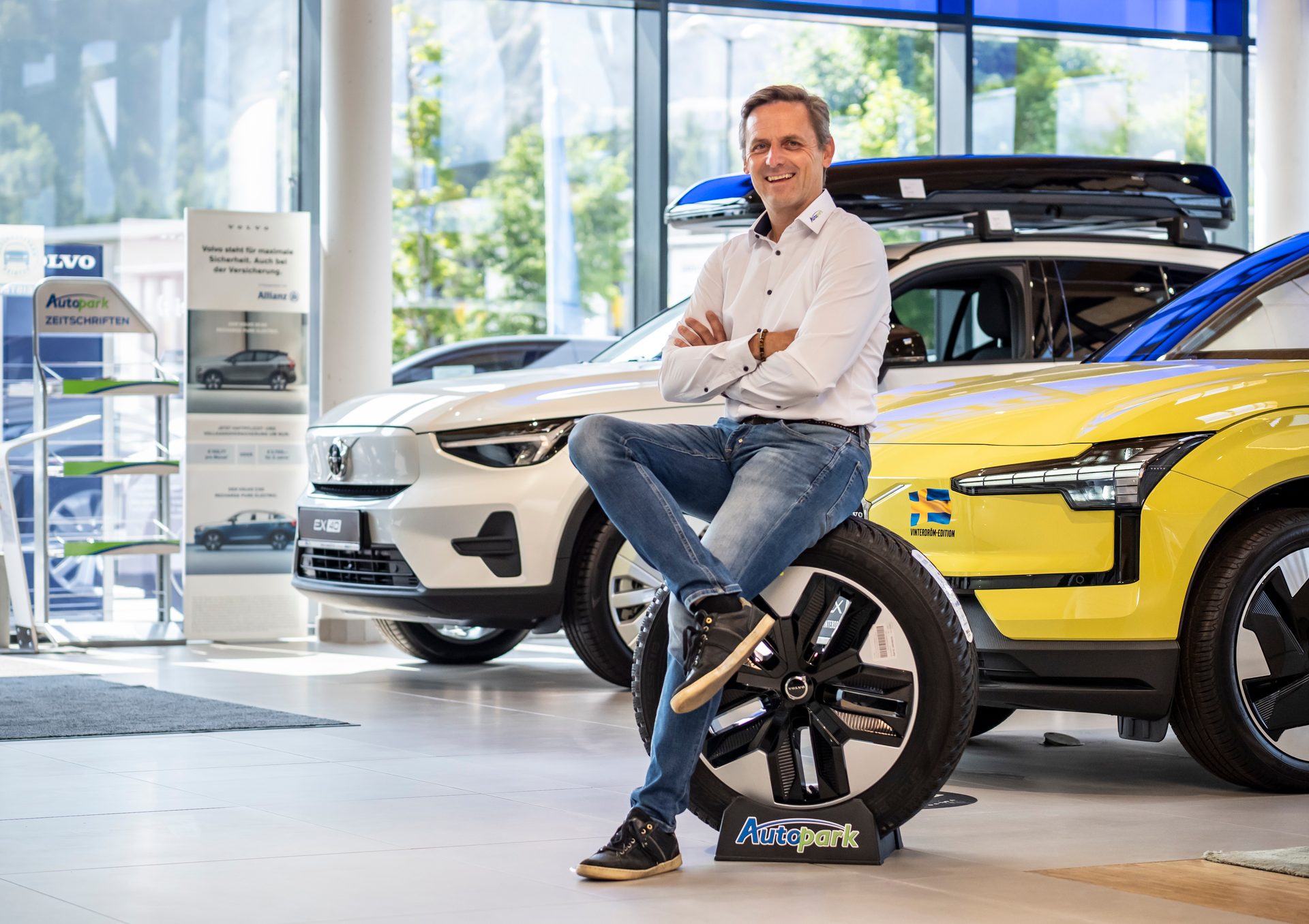 A man smiles, sitting on an "Autopark" branded tire in a Volvo car dealership with electric cars.