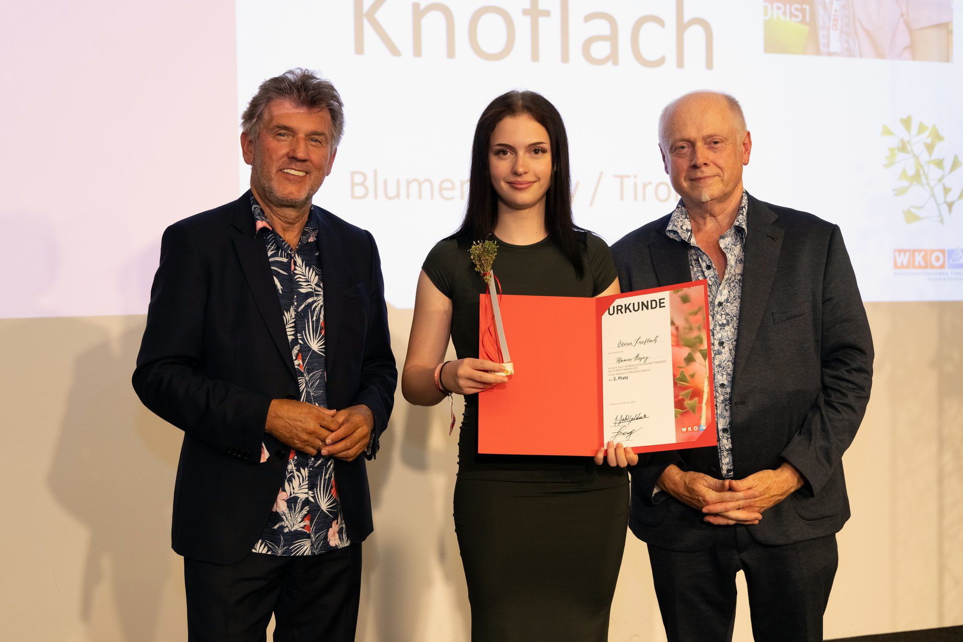 A woman holds an award, flanked by two men, on a stage.