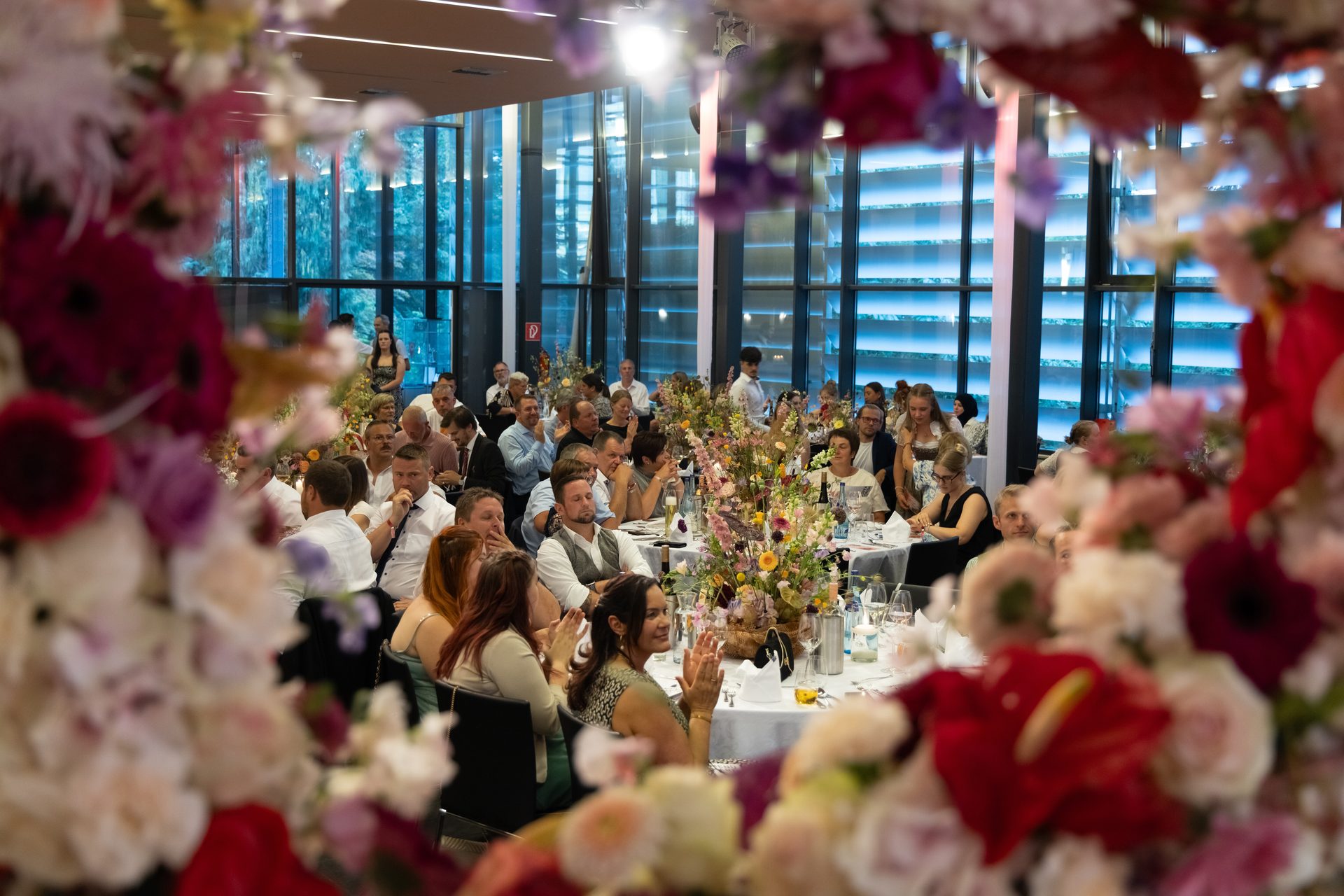 An indoor event with guests seated at tables, framed by abundant colorful flowers in the foreground.