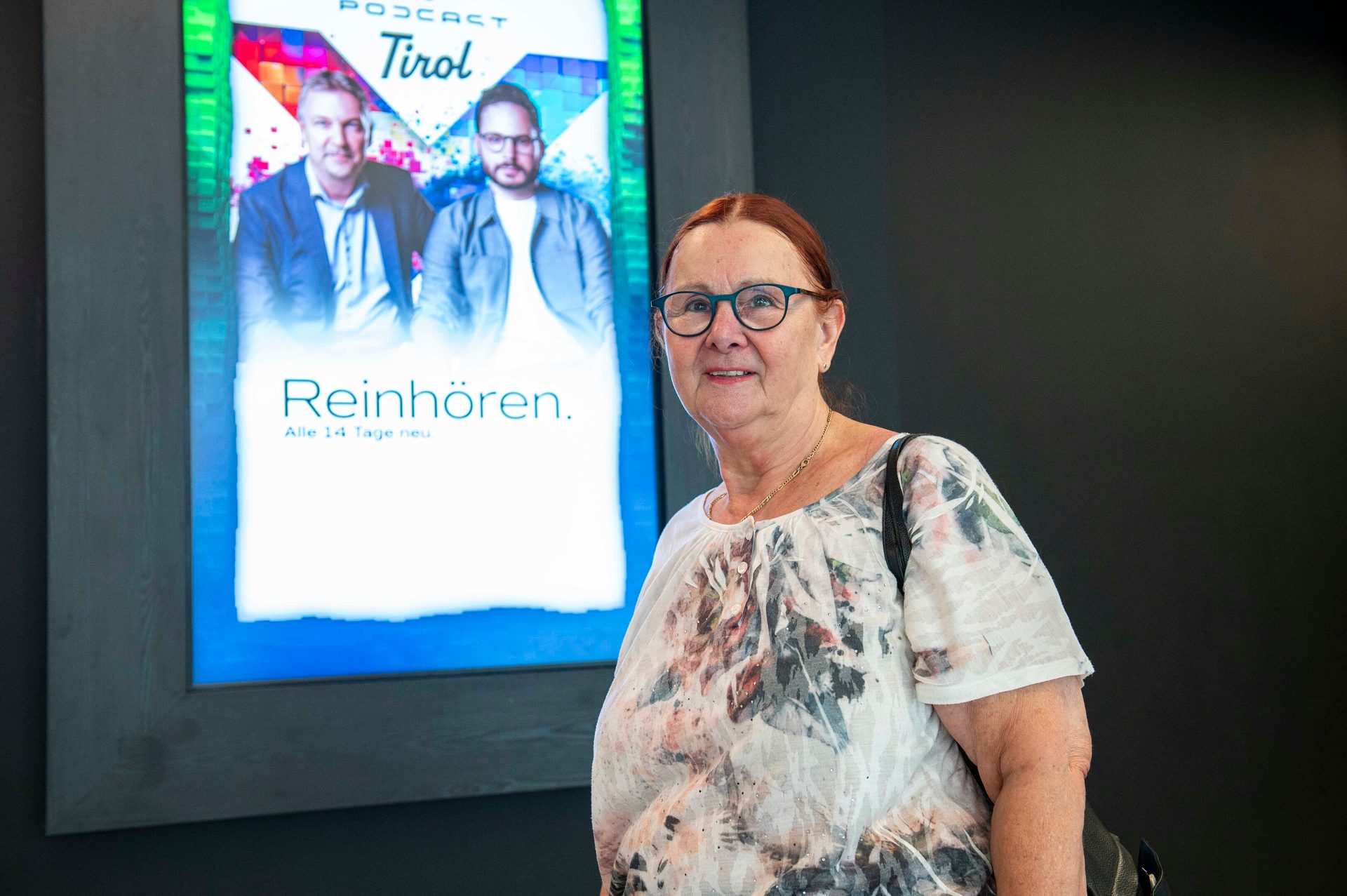 Smiling older woman in glasses stands before a digital display for "PODCAST Tirol".