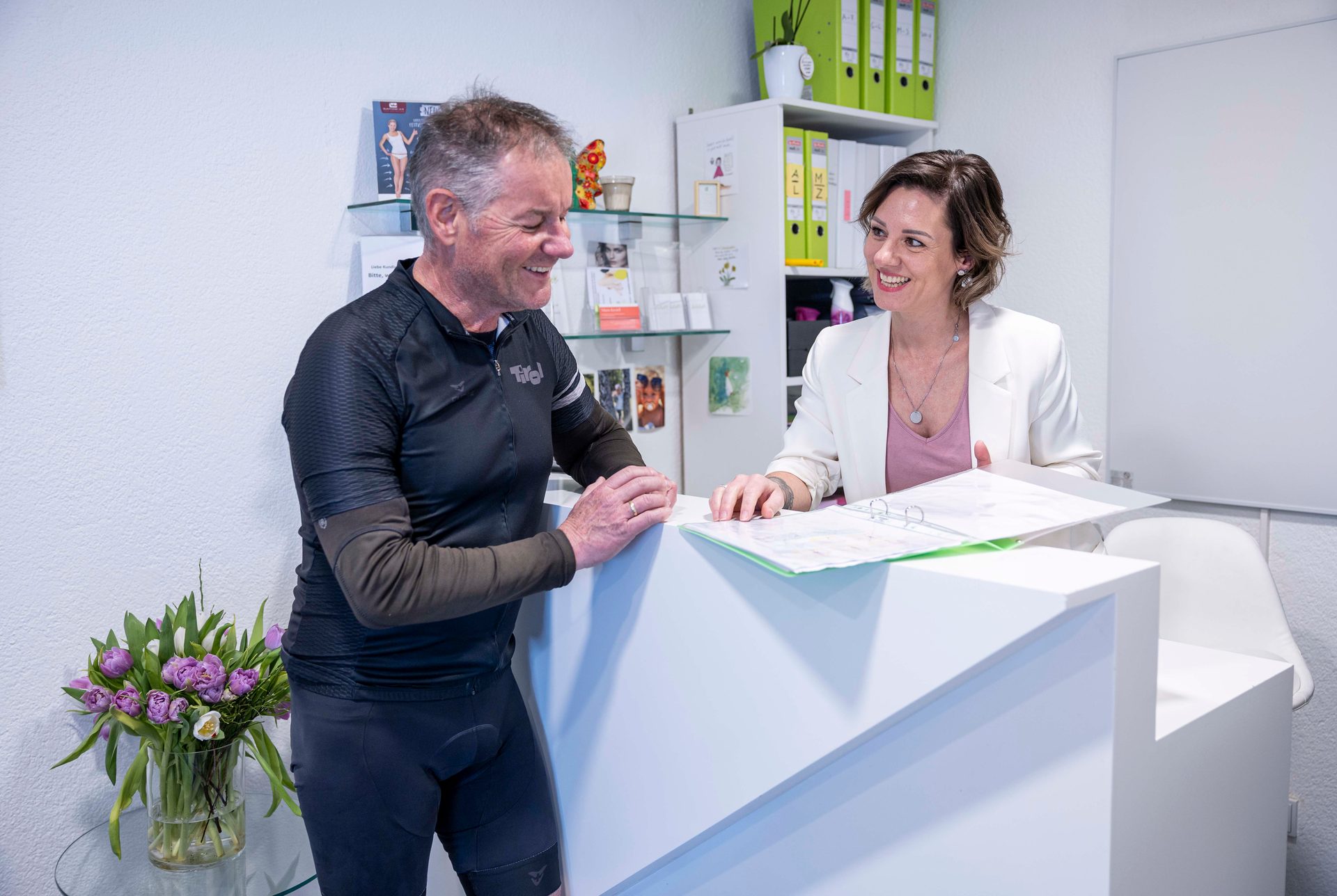 A man in cycling gear smiles at a woman holding a folder across a white reception desk.