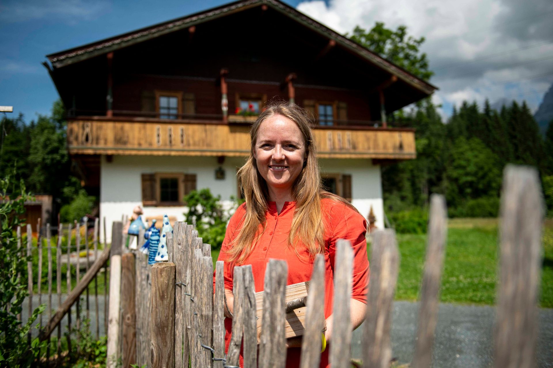 Smiling woman in red, behind a wooden fence, with a traditional house and forest behind her.