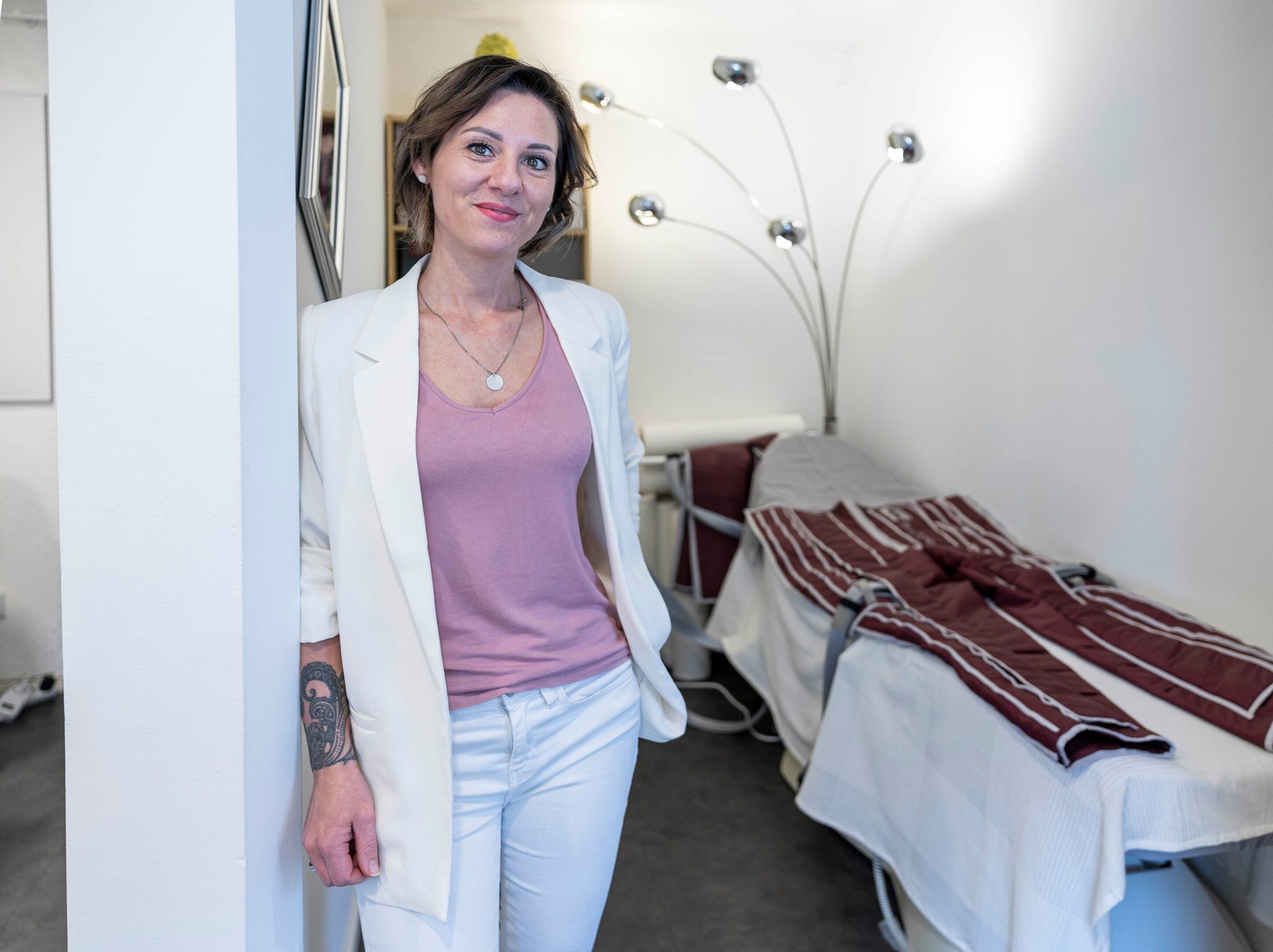 A woman in a white blazer and pink top smiles while leaning against a wall in a treatment room.