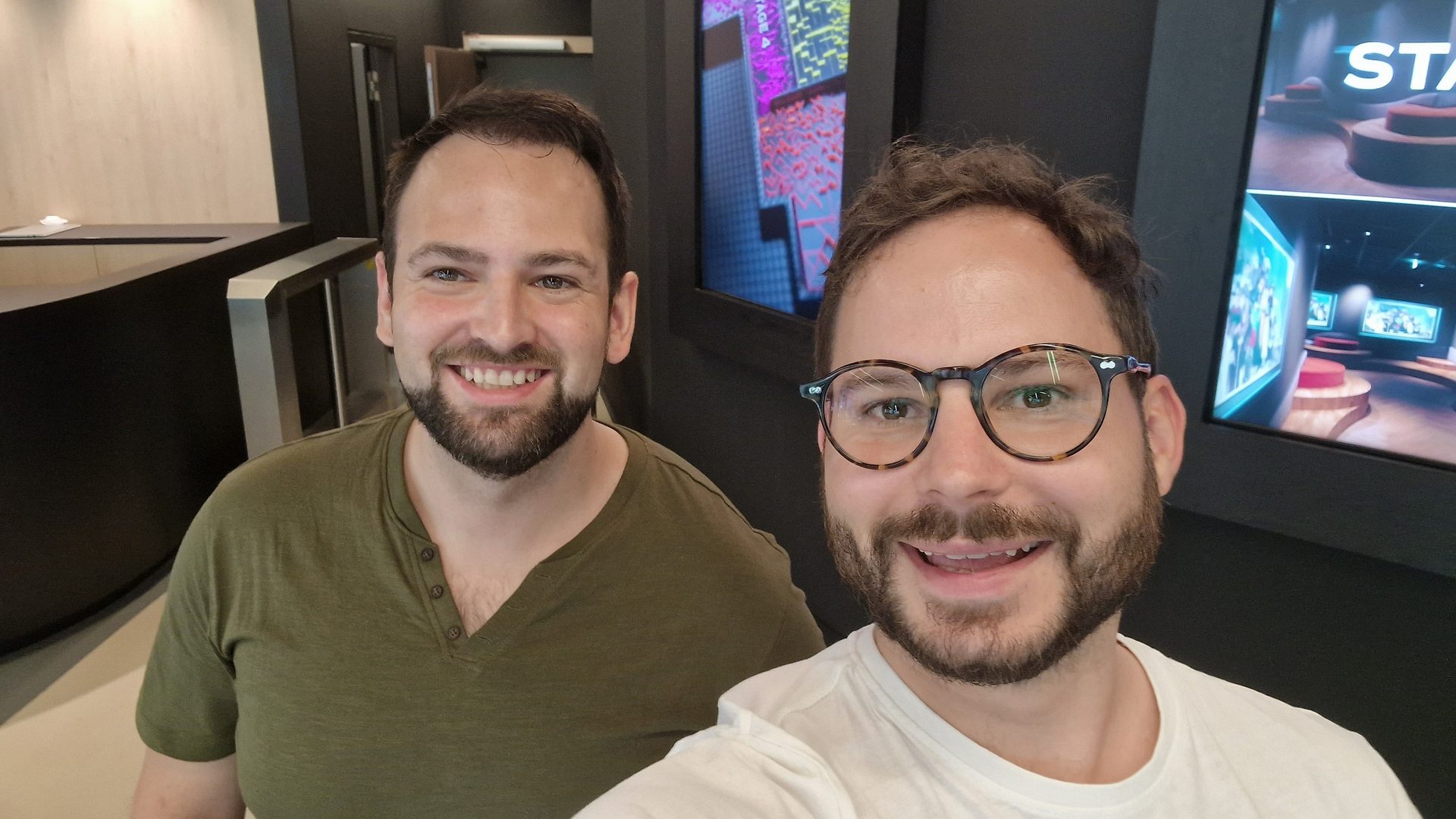 Two smiling men, one with glasses, taking a selfie indoors.