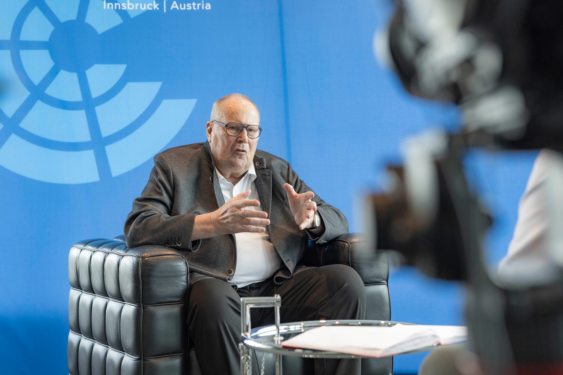 Elderly man in glasses and suit jacket seated in a chair, gesturing while speaking. A blurred camera is visible.
