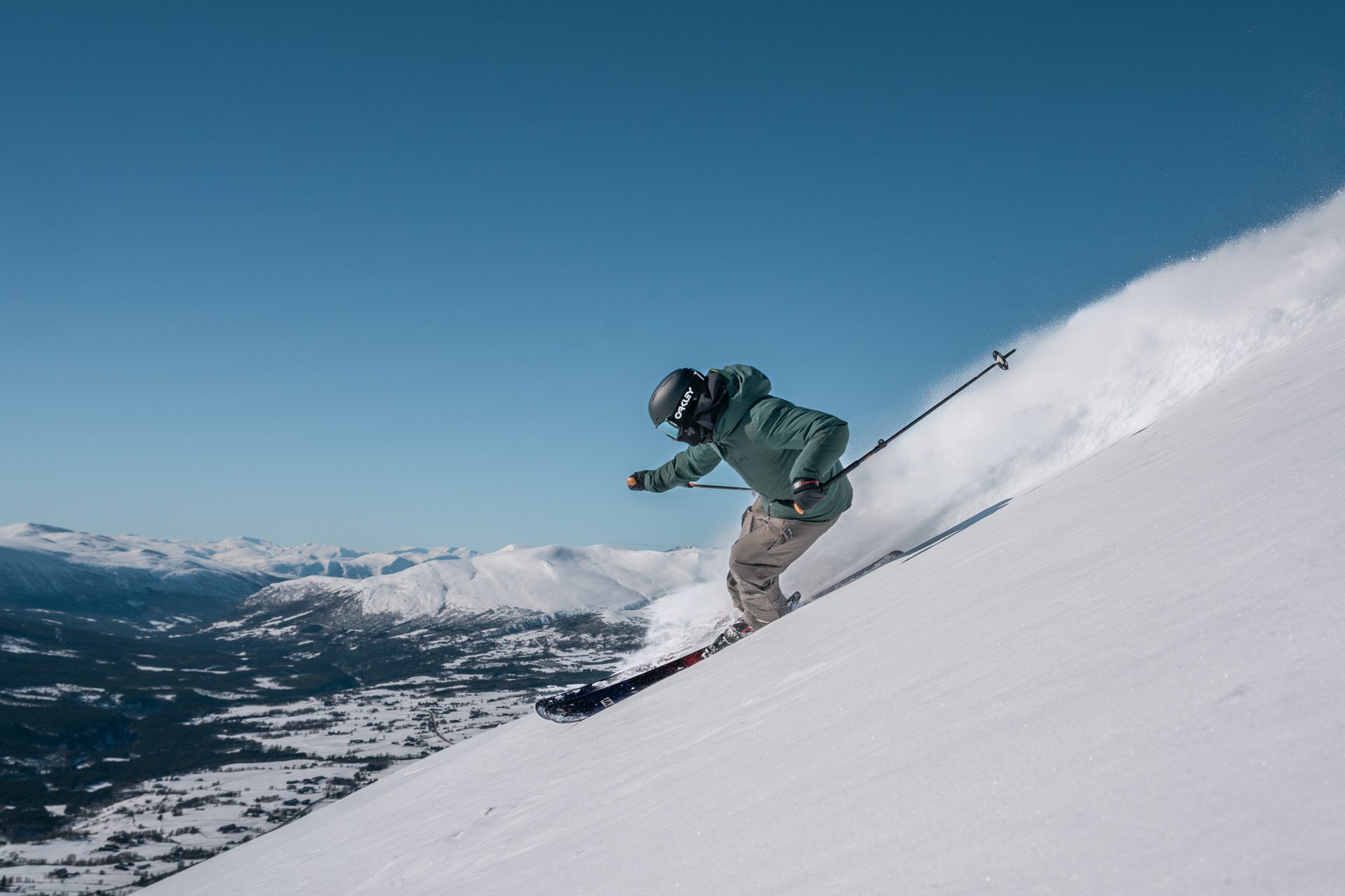 Skier in green jacket and black helmet carving on a snowy mountain with a clear blue sky.