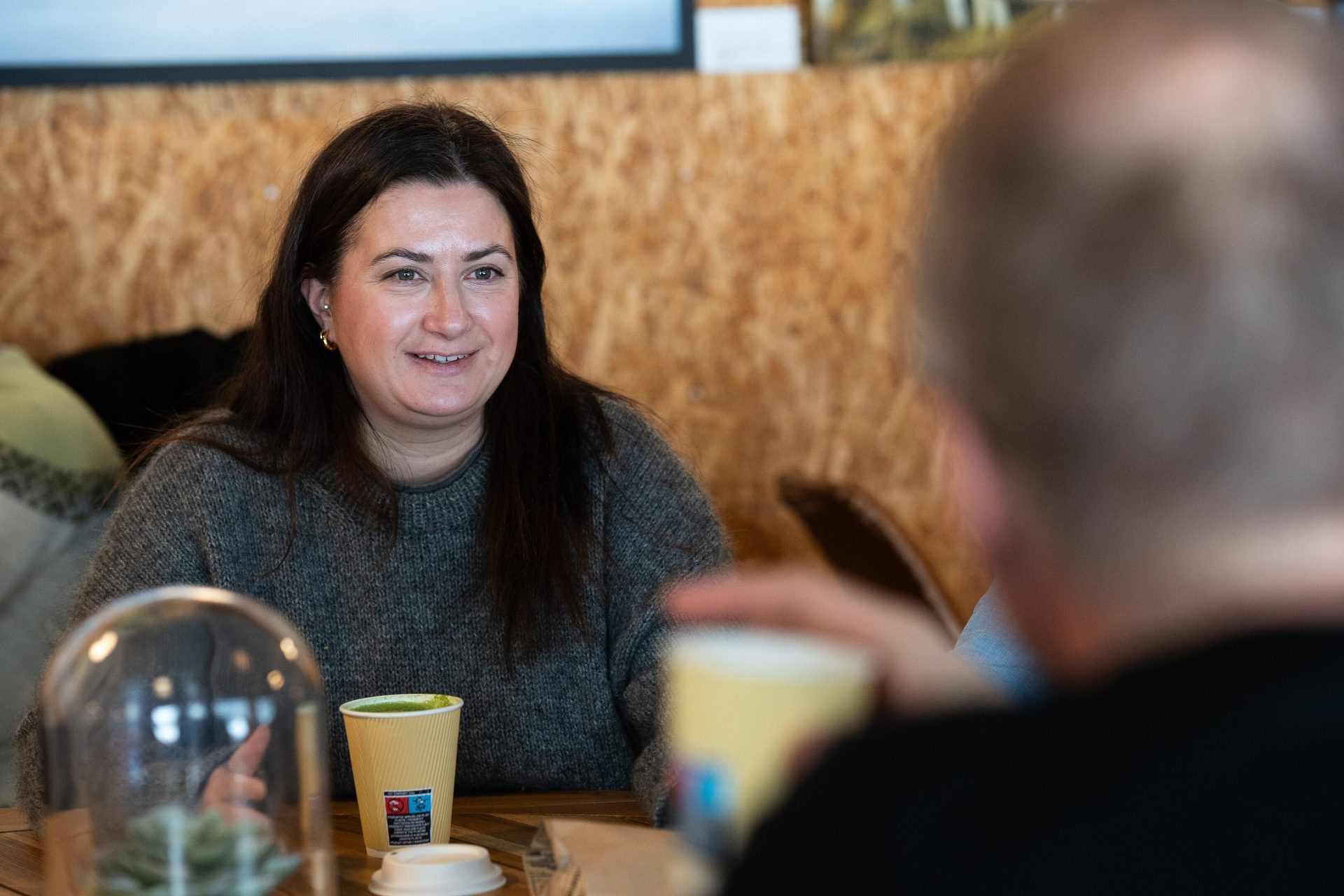 A smiling woman with dark hair sits at a cafe table with a green drink, conversing with someone.