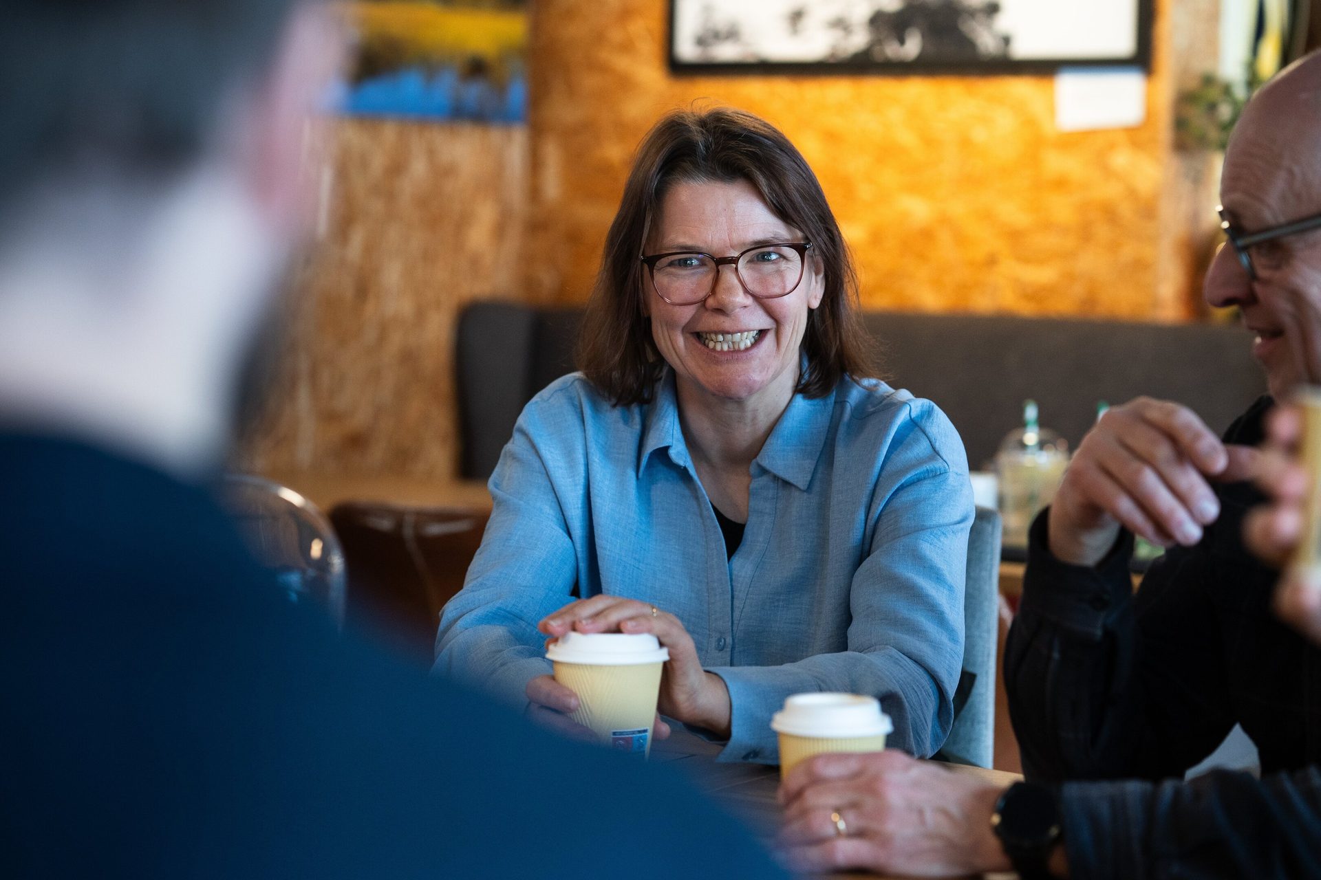 A smiling woman in glasses sits at a table with two men, holding coffee cups.