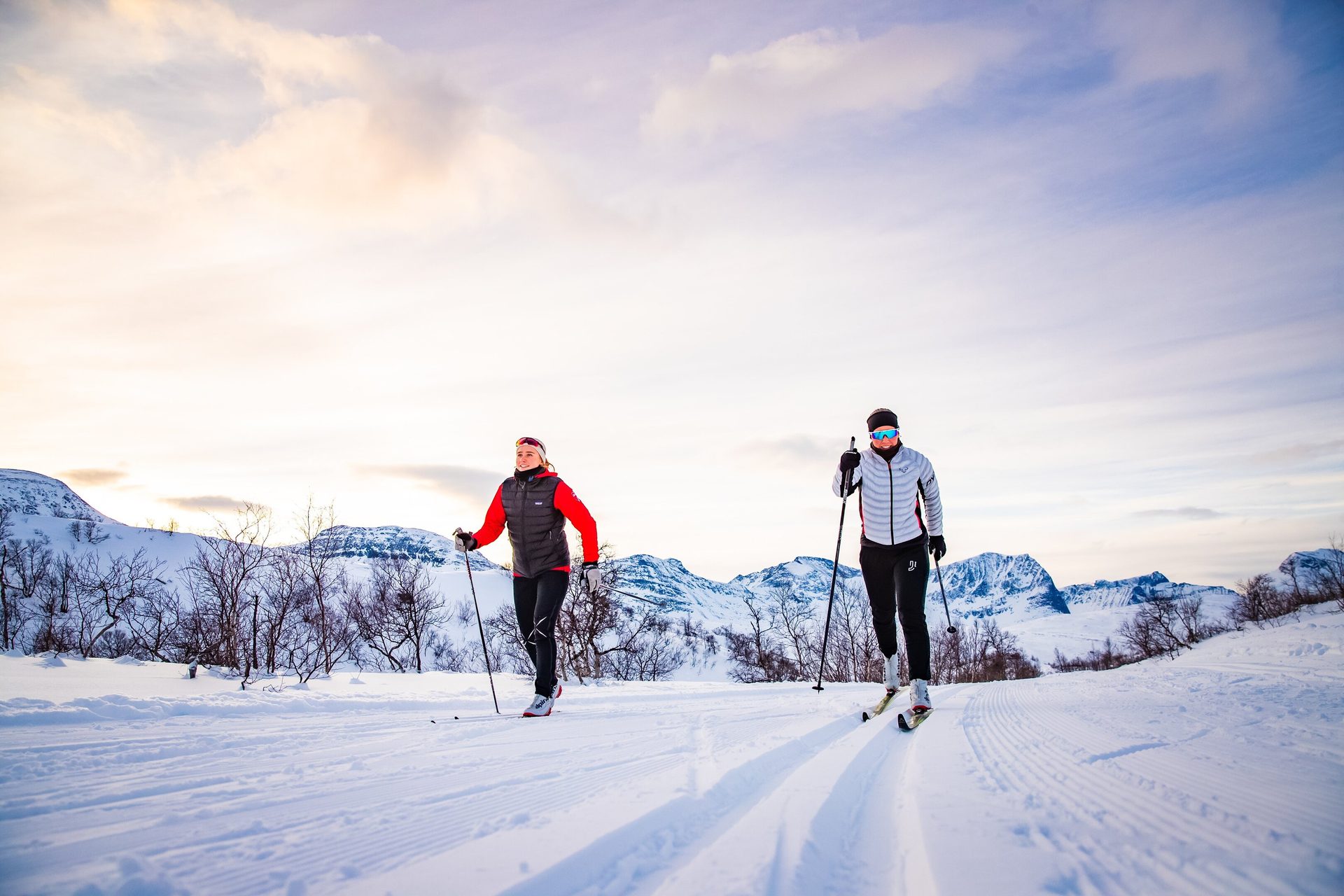 Two people cross-country skiing on a snowy track with mountains and a bright sky in the background.