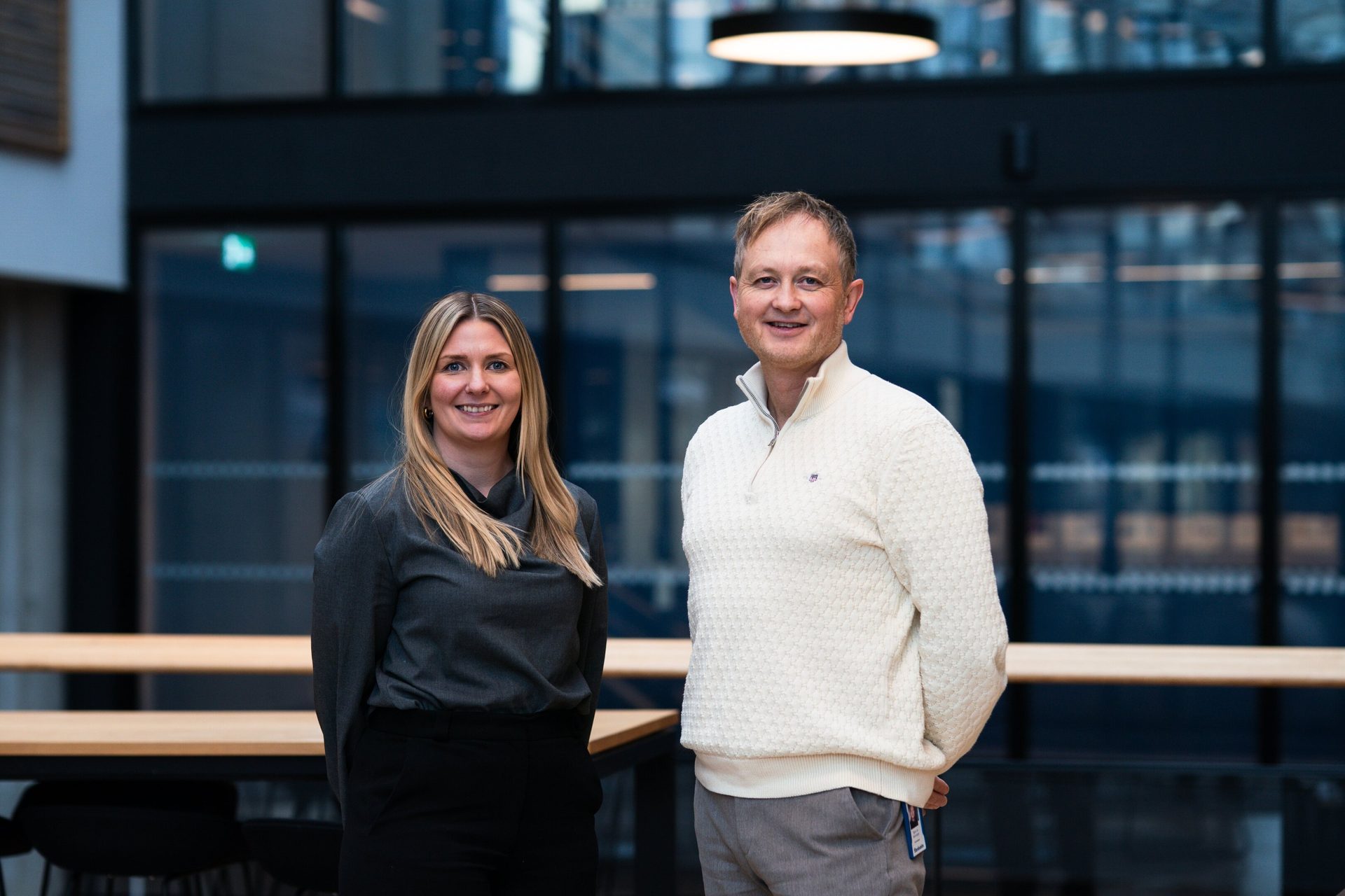 Two smiling professionals, a woman and a man, standing together in a modern office environment.
