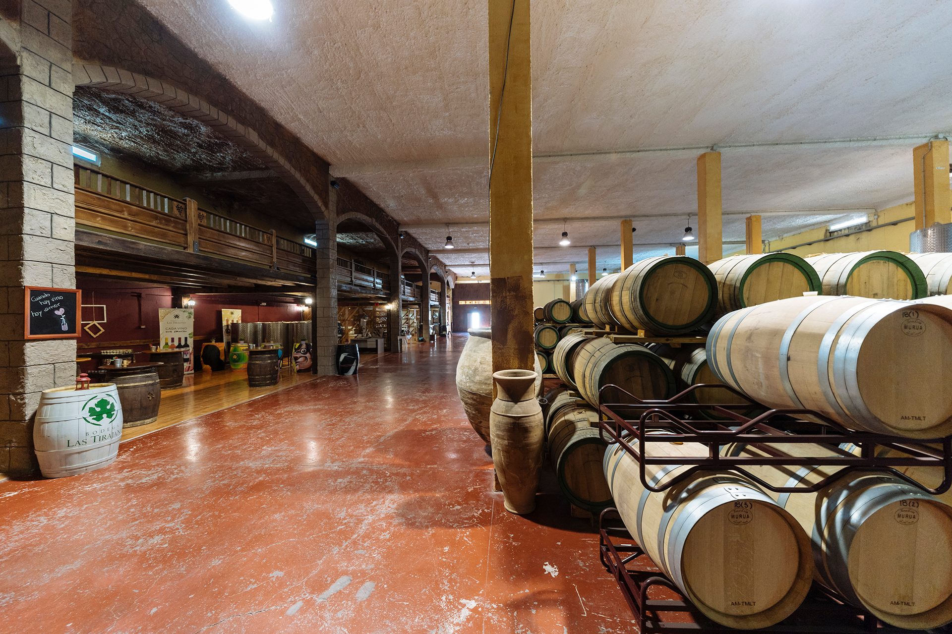 A wine cellar with rows of stacked oak barrels, a red floor, stone pillars, and a dark wooden mezzanine.