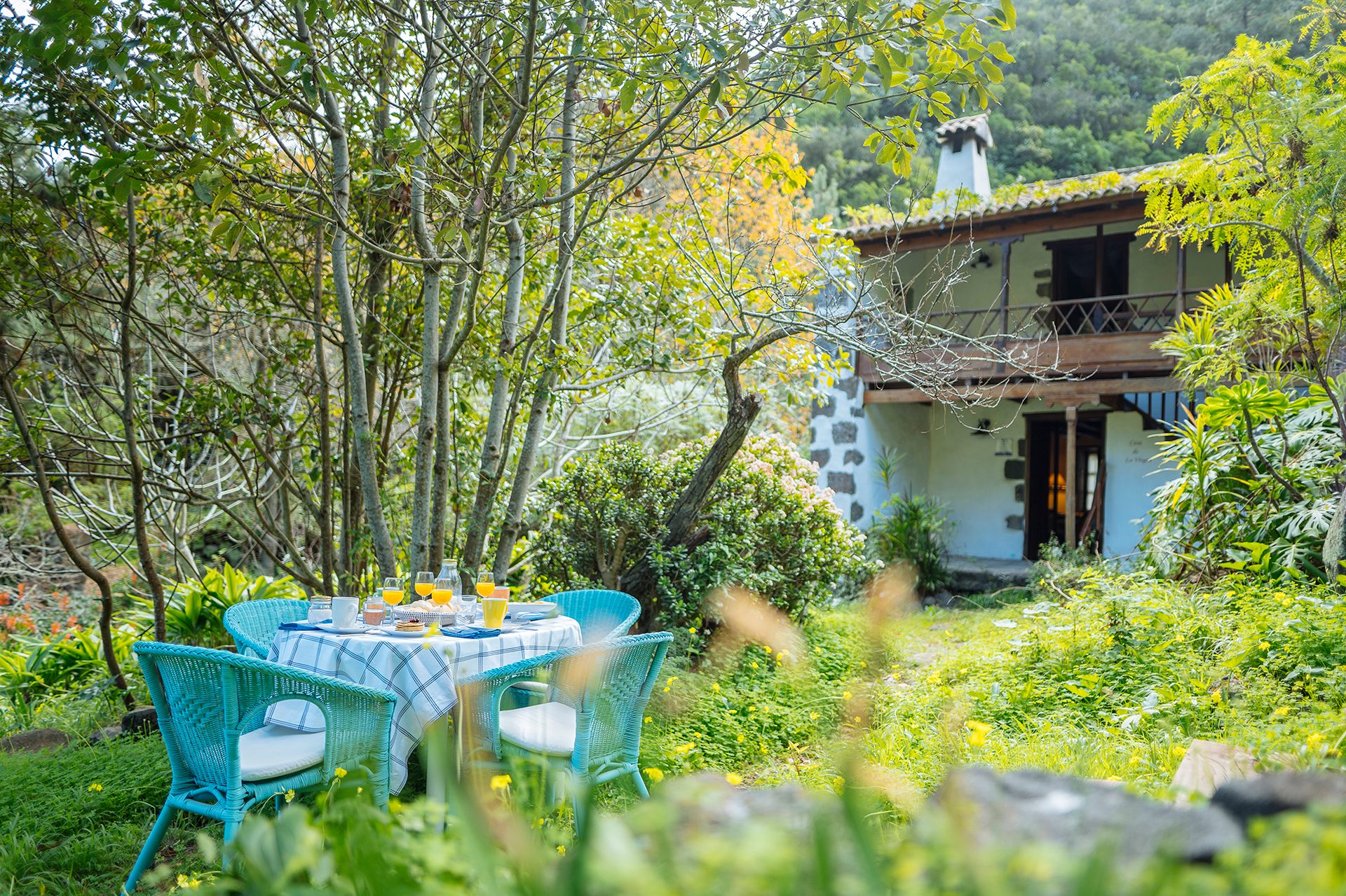 Outdoor breakfast table with turquoise chairs in a lush garden, with a rustic house behind.