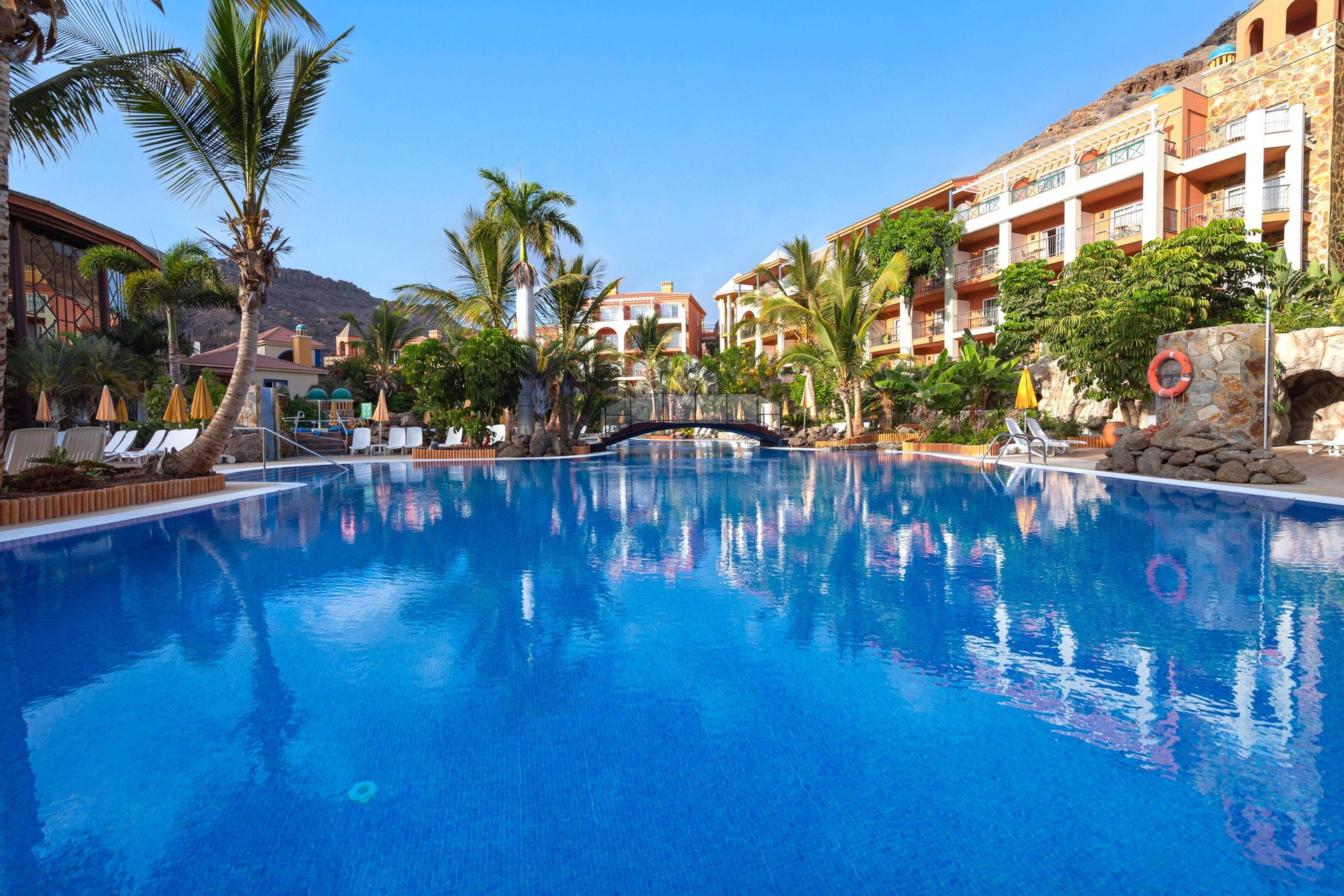 Resort pool with blue water, palm trees, hotel buildings, and a small bridge under a clear sky.