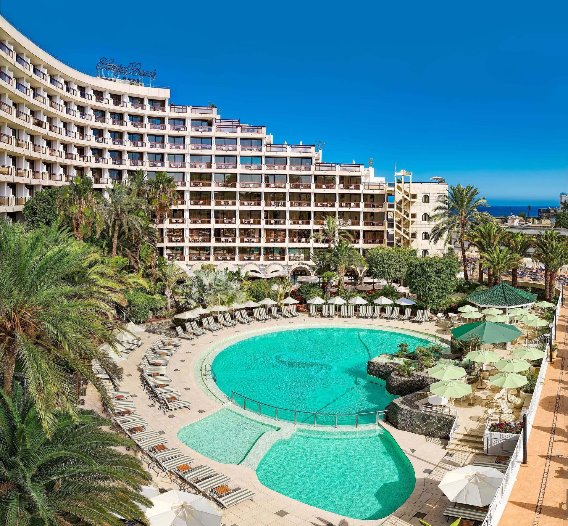 Curved hotel, pool with loungers & green umbrellas, palm trees, blue sky, distant ocean.