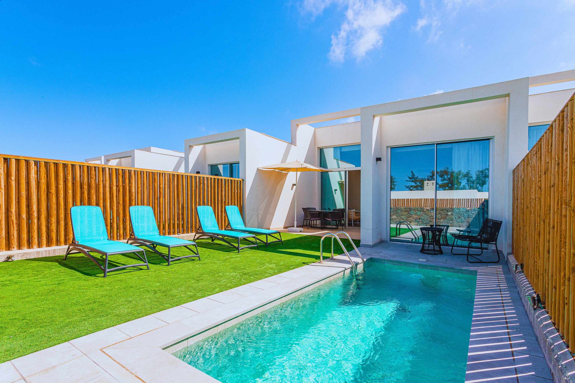 Modern villa with a private pool, artificial turf, lounge chairs, and a wooden fence under a blue sky.