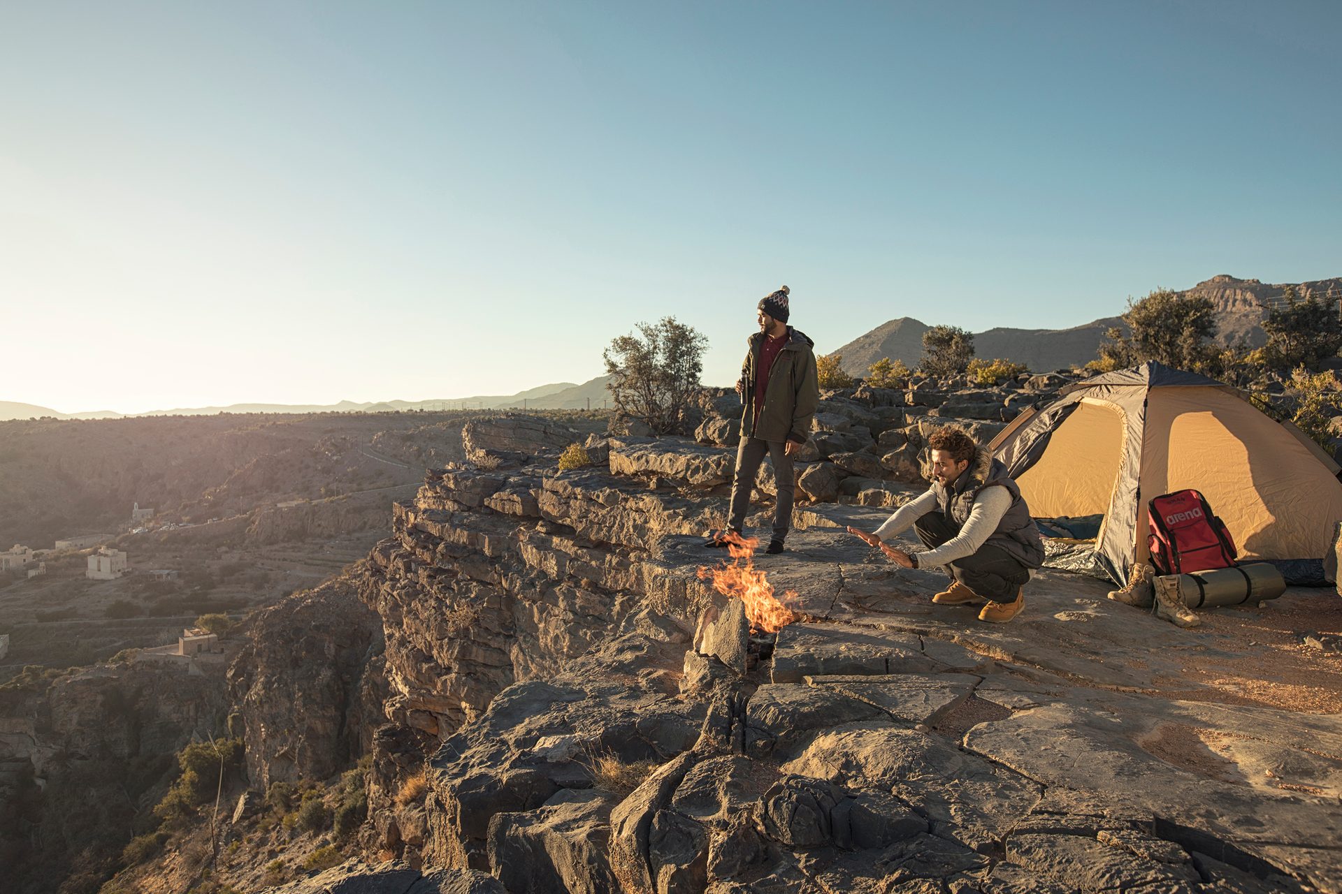 Two men camp on a rocky cliff by a fire, with a tent and vast mountain views.