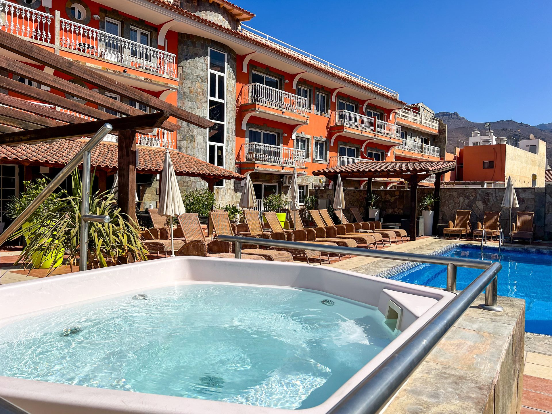 Vibrant orange hotel with balconies, jacuzzi in foreground, swimming pool, and mountains.
