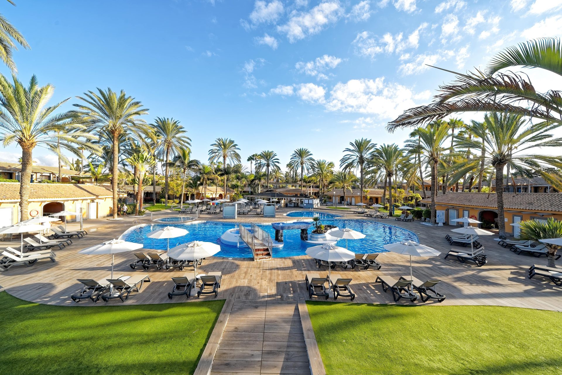 Resort pool surrounded by palm trees, sun loungers, and buildings under a blue sky.