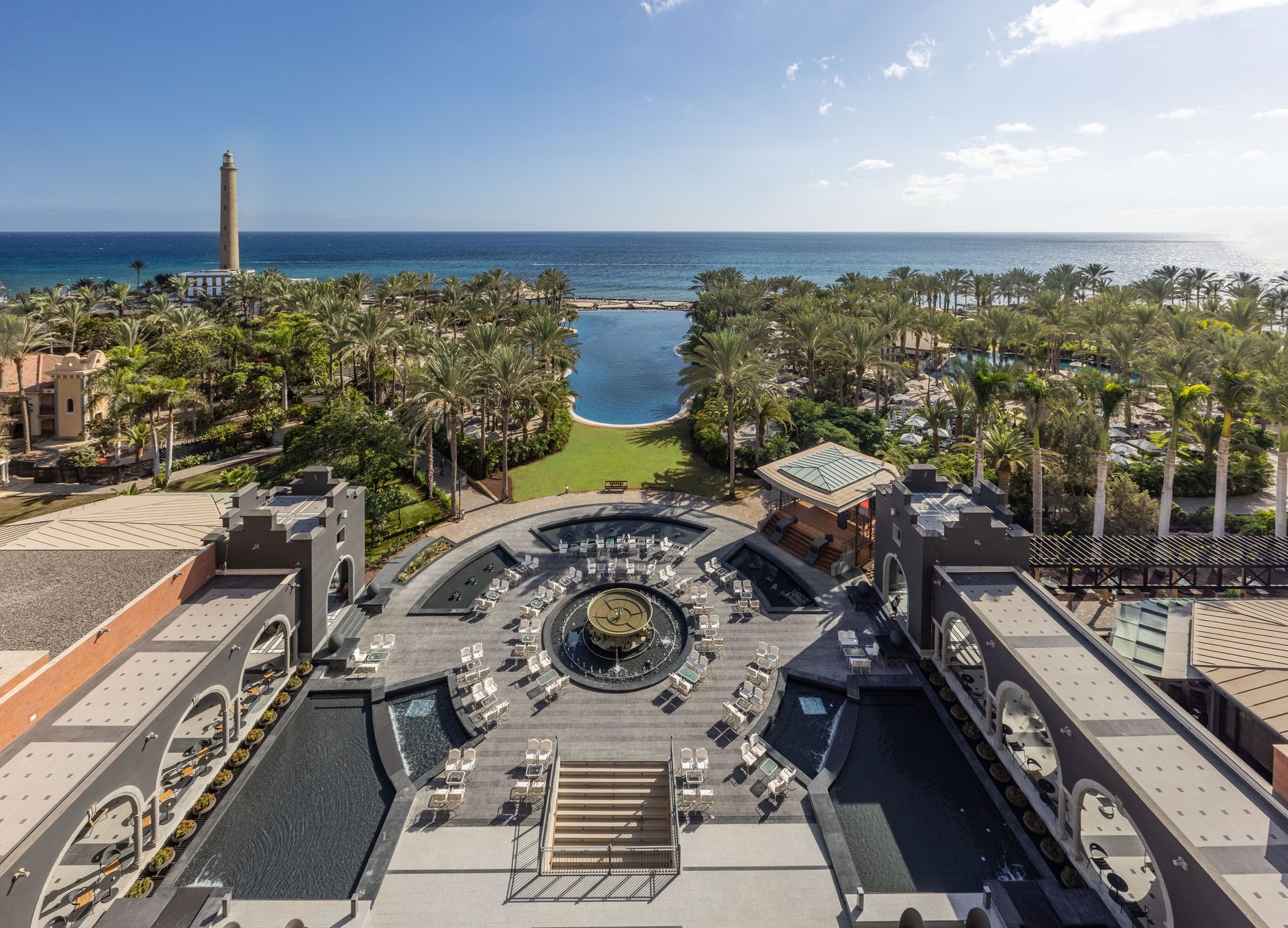 Aerial view of a resort with patio, palm trees, infinity pool, ocean, and lighthouse.