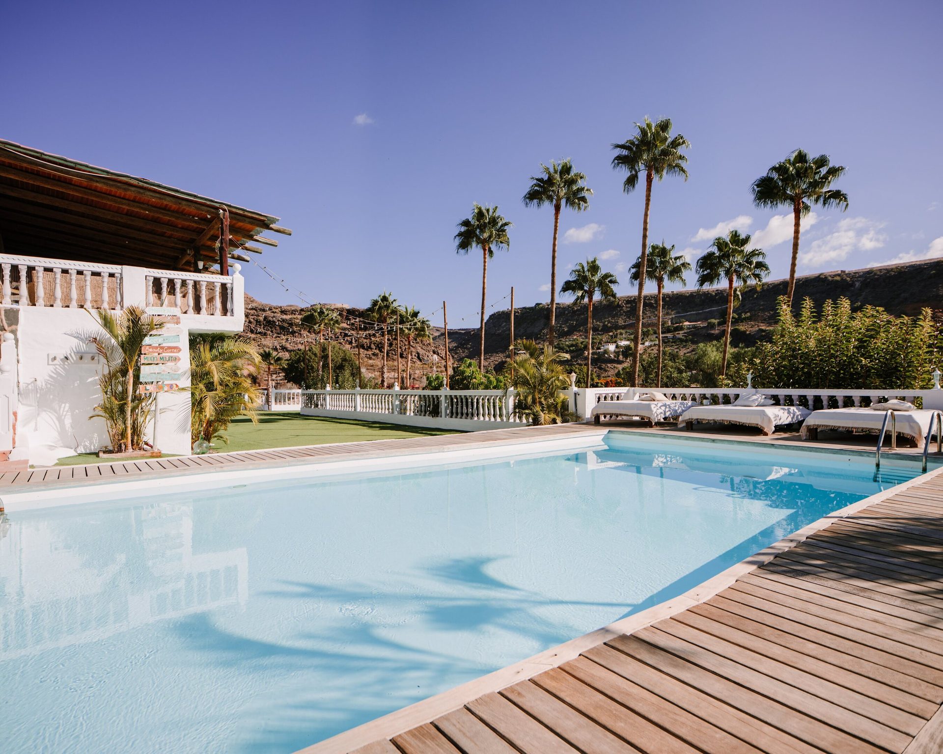 Sunny pool with a white building, palm trees, sun loungers, and a wooden deck.