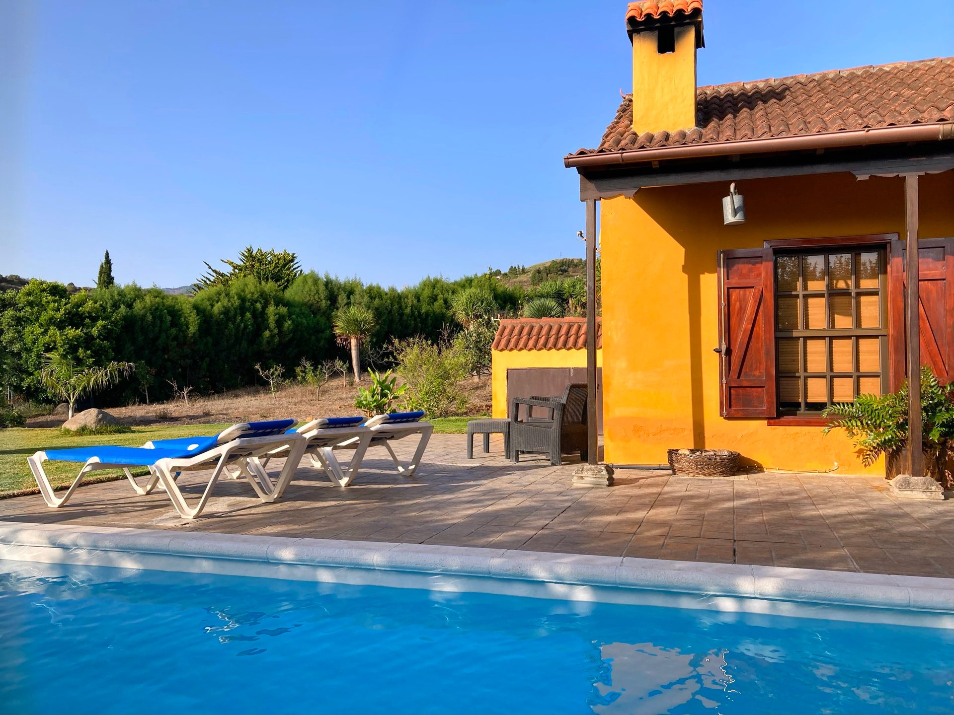 Yellow house with a tiled roof, blue swimming pool, lounge chairs, and green trees under a clear sky.